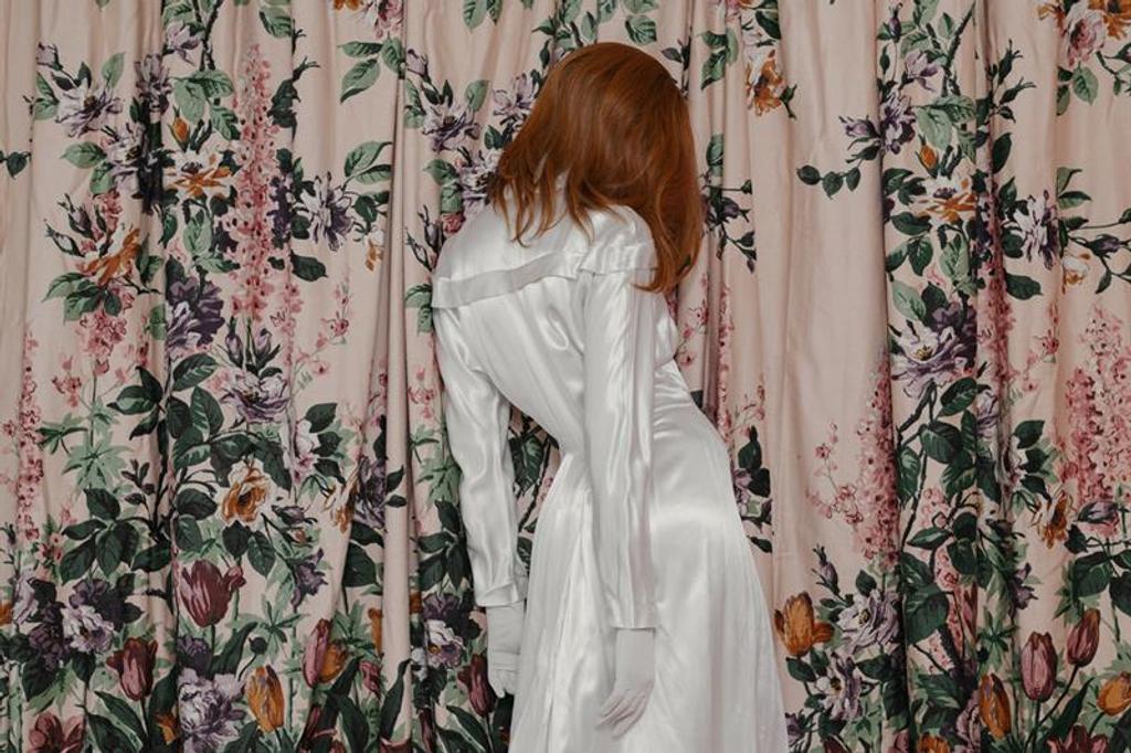 A woman in a white dress stands in front of a pink curtain decorated with flowers.