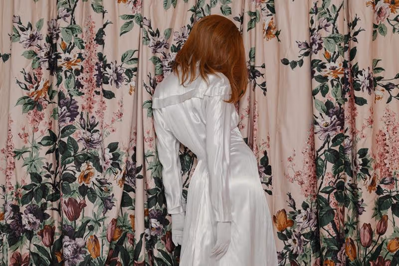 A woman in a white dress stands in front of a pink curtain decorated with flowers.