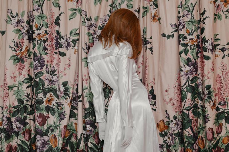 A woman in a white dress stands in front of a pink curtain decorated with flowers.