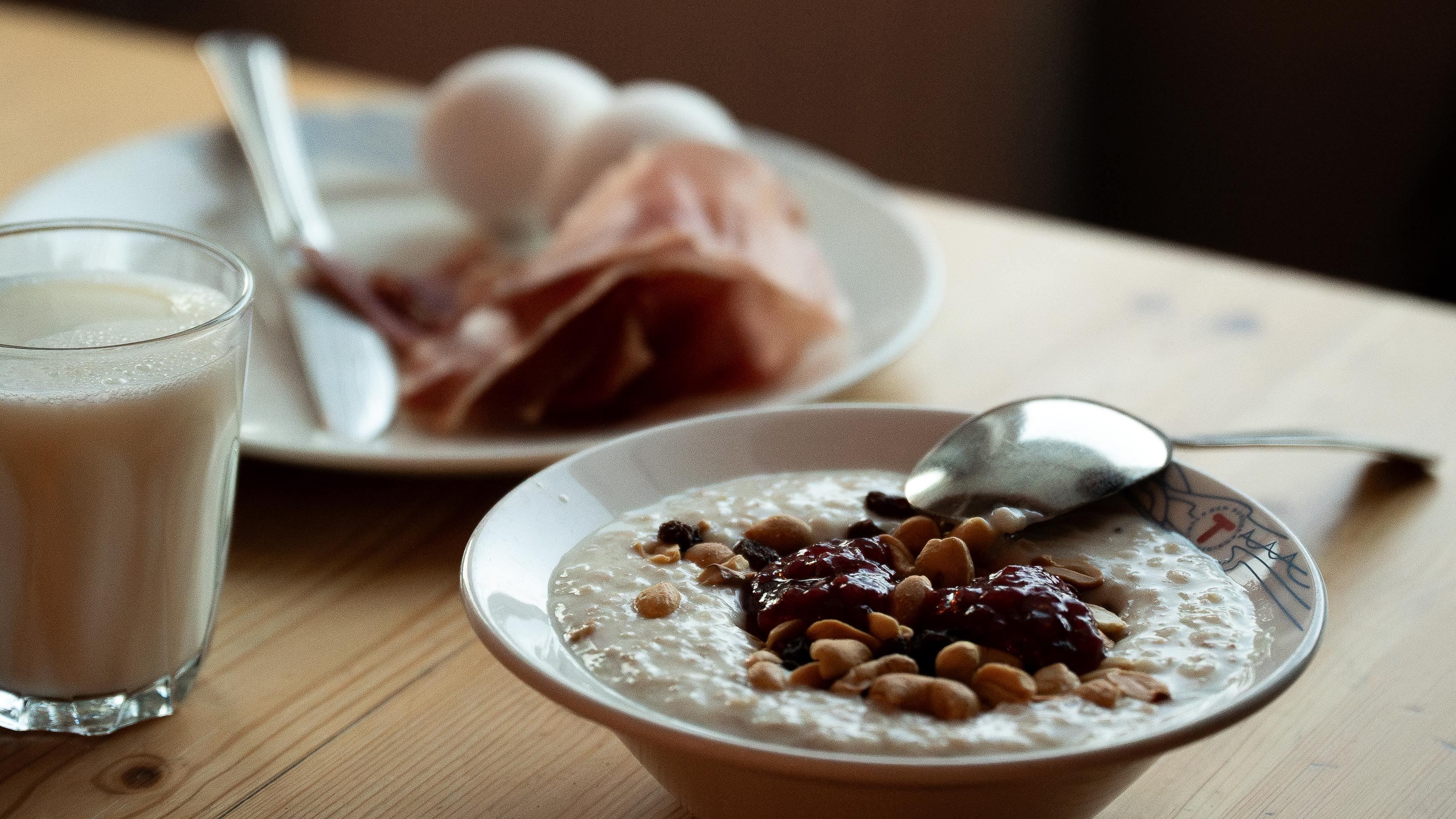 Bowl of porridge topped with jam and nuts on a wooden table, with a spoon, milk glass, and eggs in the background..