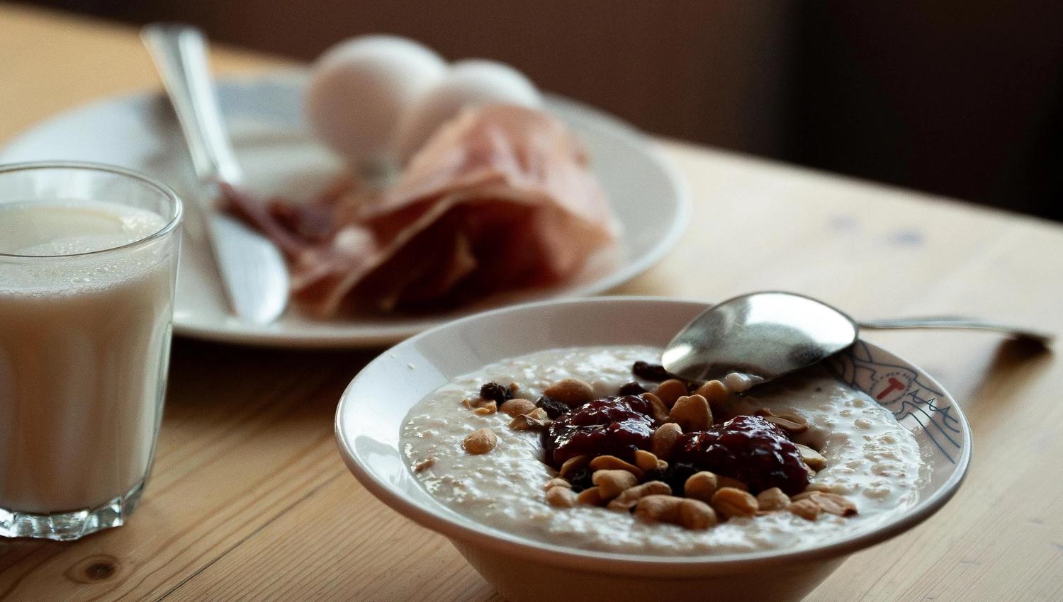 Bowl of porridge topped with jam and nuts on a wooden table, with a spoon, milk glass, and eggs in the background..