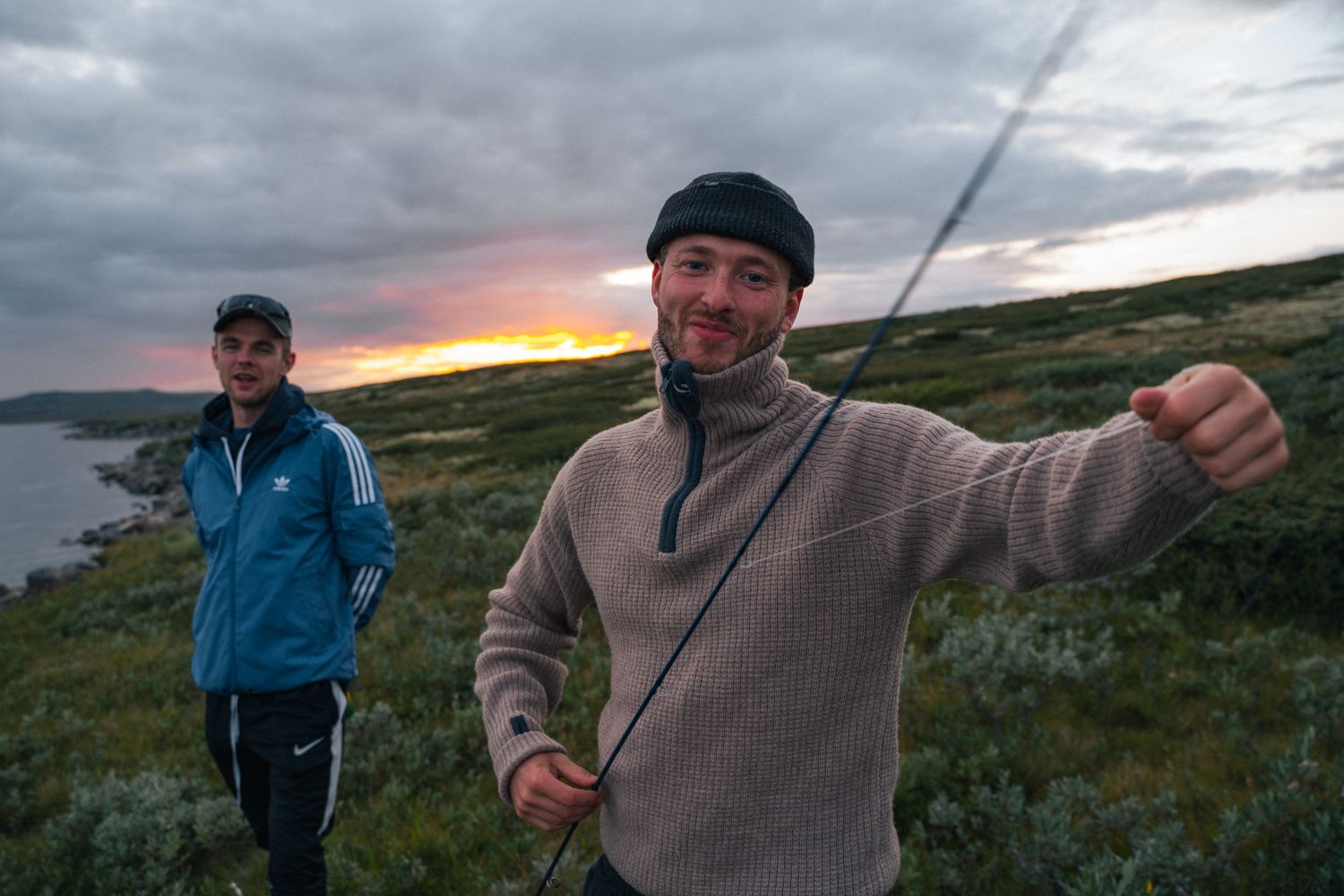 Two smiling men by a mountain lake, one of them holding a fishing rod.