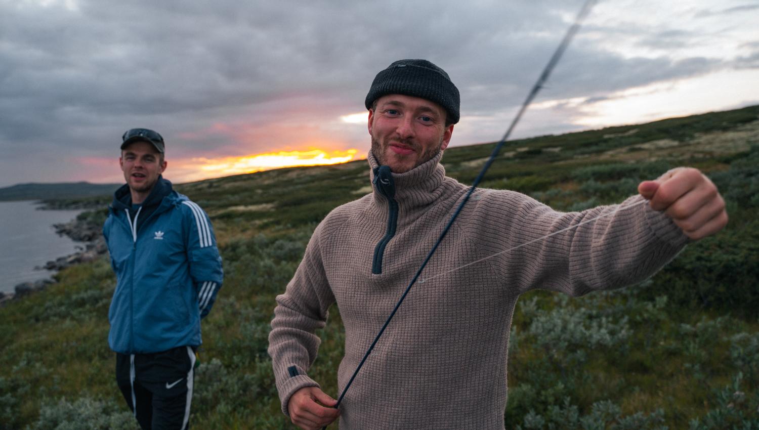 Two smiling men by a mountain lake, one of them holding a fishing rod.