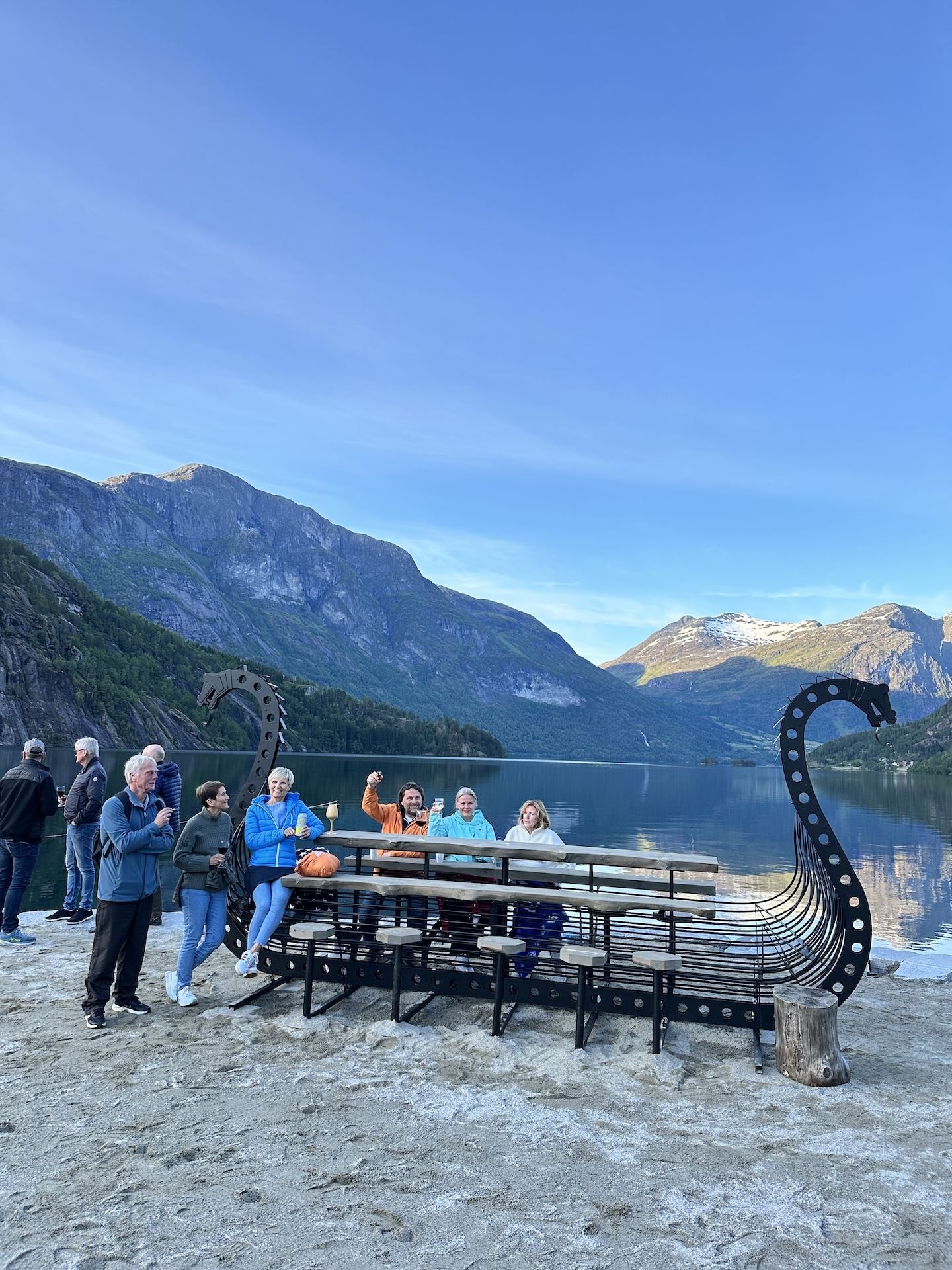 Group of people by a dragon-prow bench on a sandy lakeshore with calm water and mountains under blue sky..