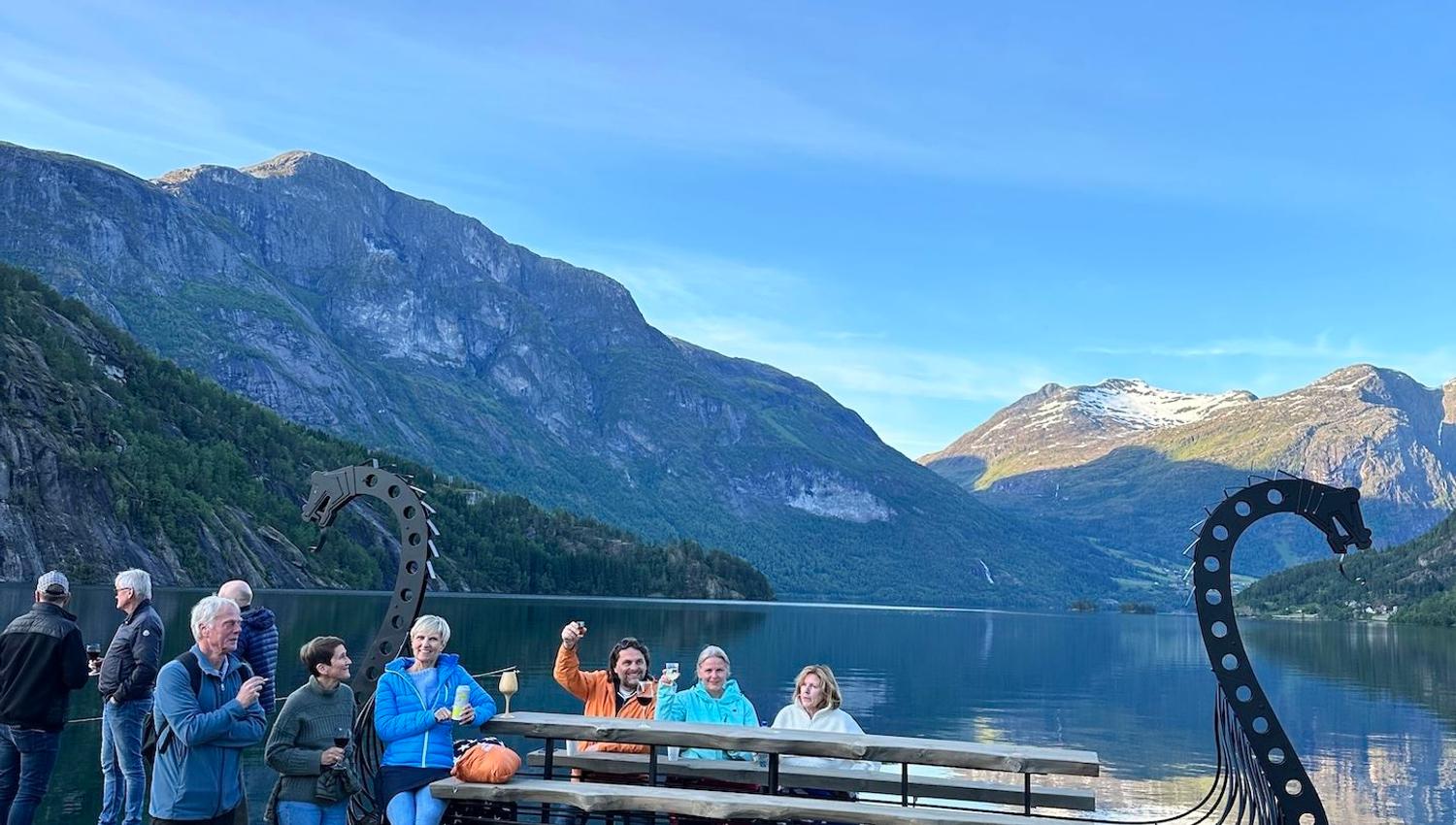 Group of people by a dragon-prow bench on a sandy lakeshore with calm water and mountains under blue sky..