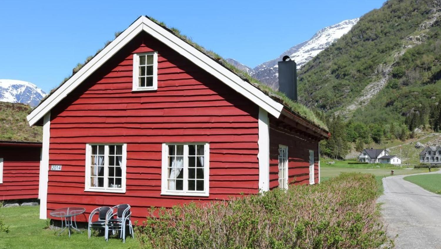 Red wooden cabin with white-trim windows and grass roof beside a lawn, road, and snowy mountains under blue sky at Aabrekk Gard Trollbu in Oldedalen valley.