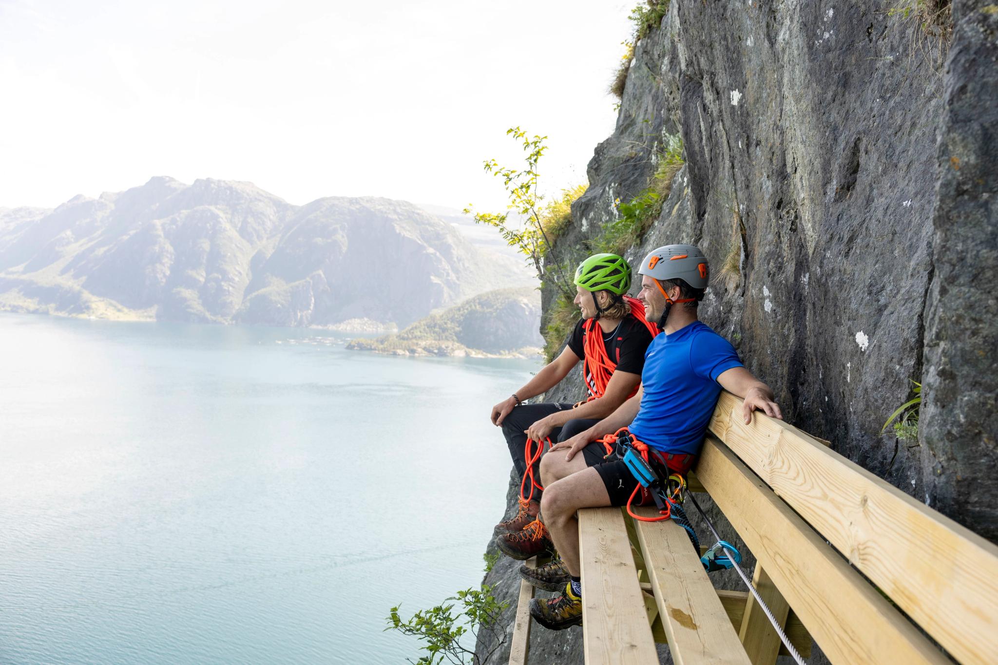 Two helmeted climbers sit on a wooden ledge beside a rock wall, overlooking a calm fjord and distant mountains.