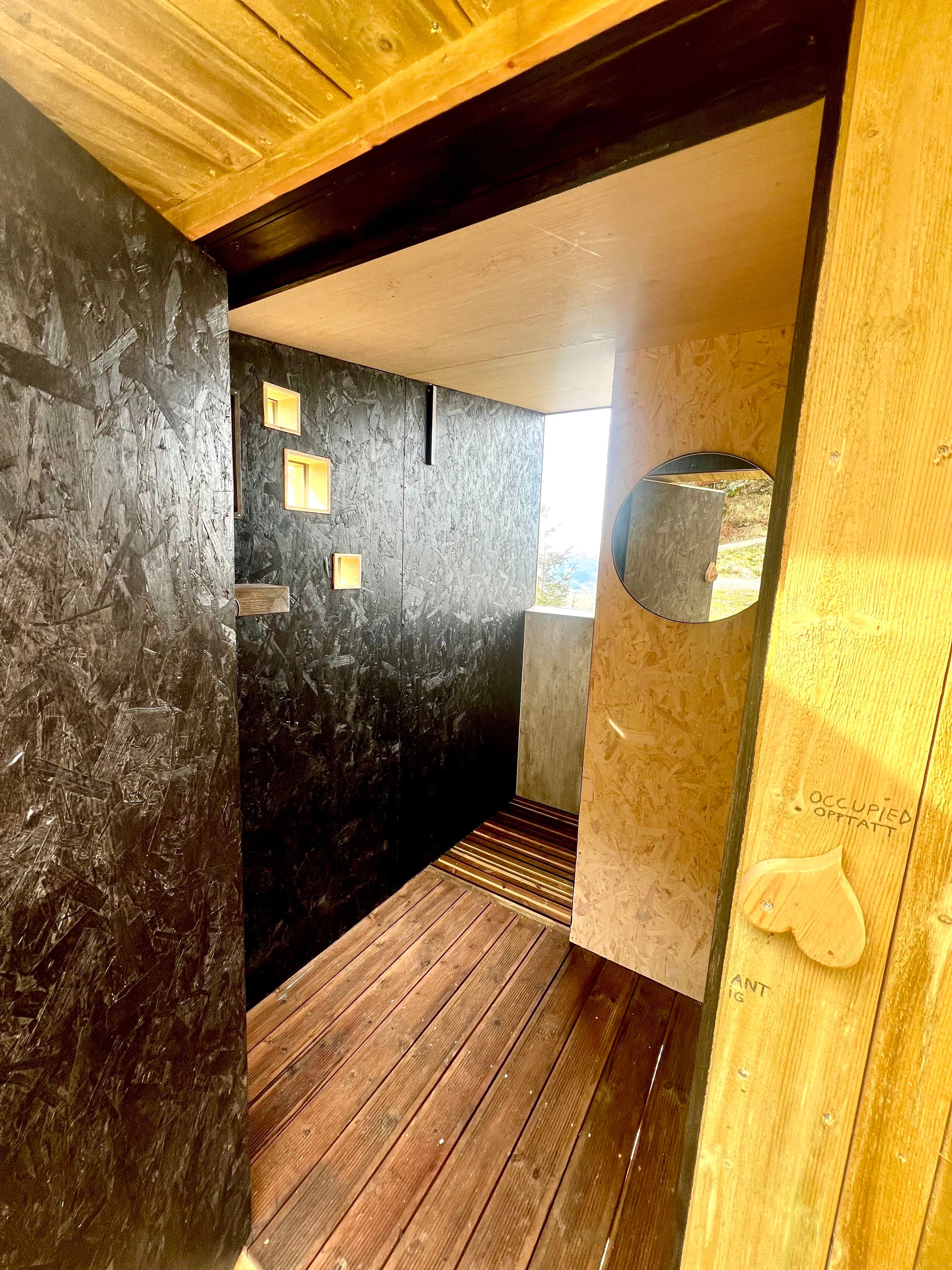 Rustic outdoor shower stall with dark walls, wooden floor, small square windows, and round mirror near doorway..
