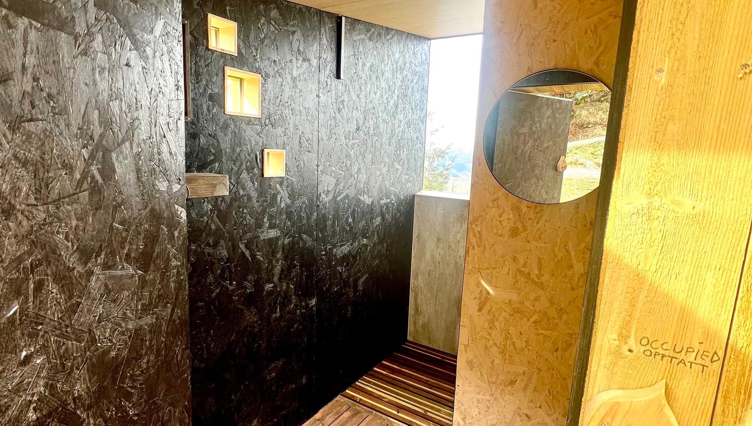 Rustic outdoor shower stall with dark walls, wooden floor, small square windows, and round mirror near doorway..
