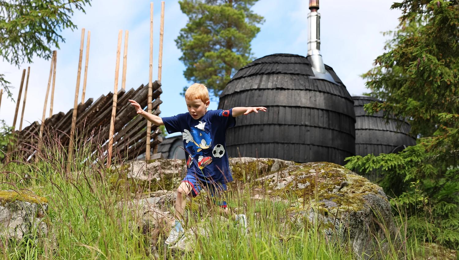 Barn som leker i gresset ved en tønneformet grillhytte i skogsomgivelser.
