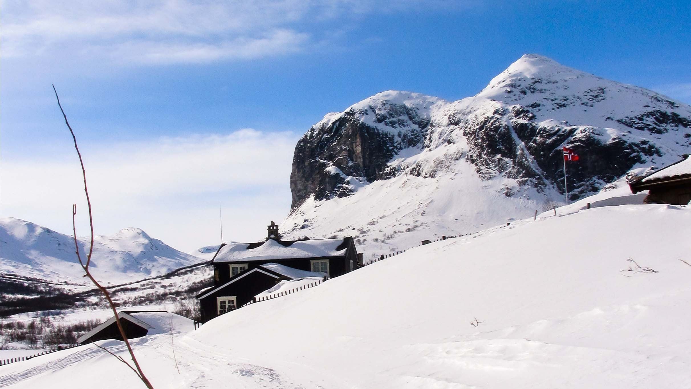 Snow-covered cabins sit in a winter valley below a rugged mountain peak, under a bright blue sky with light clouds..