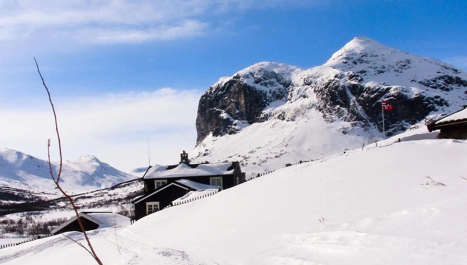 Snow-covered cabins sit in a winter valley below a rugged mountain peak, under a bright blue sky with light clouds..
