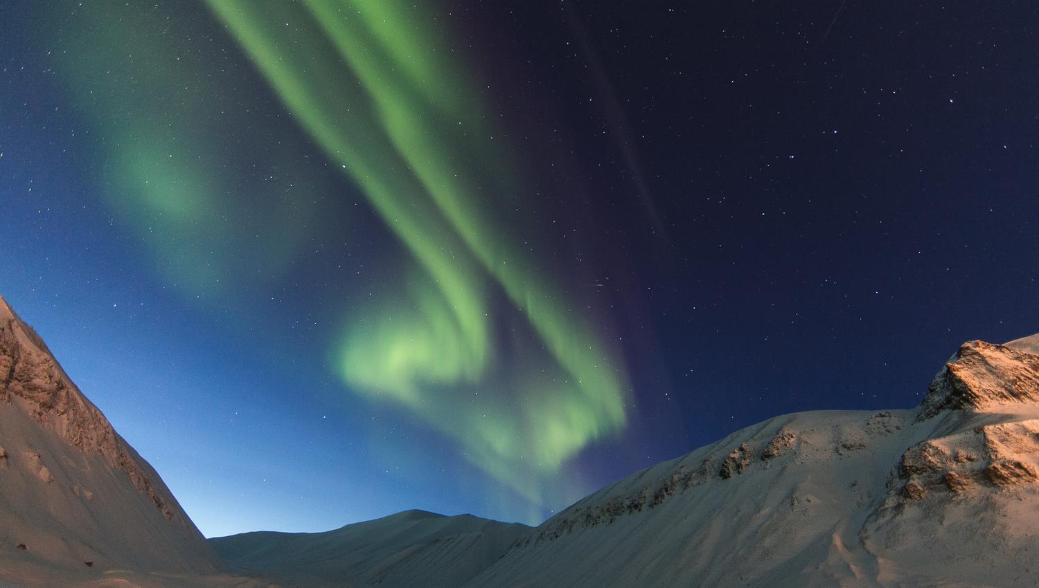 A mountainous landscape in the foreground with northern lights in the skies in the background