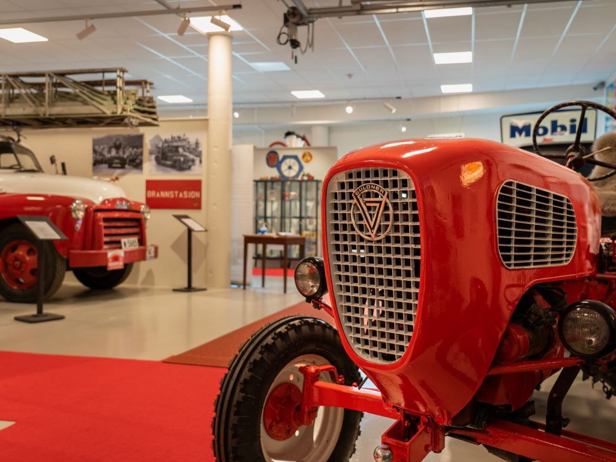 Close-up of a red vintage tractor front in an indoor museum, with another tractor and exhibits in the background.