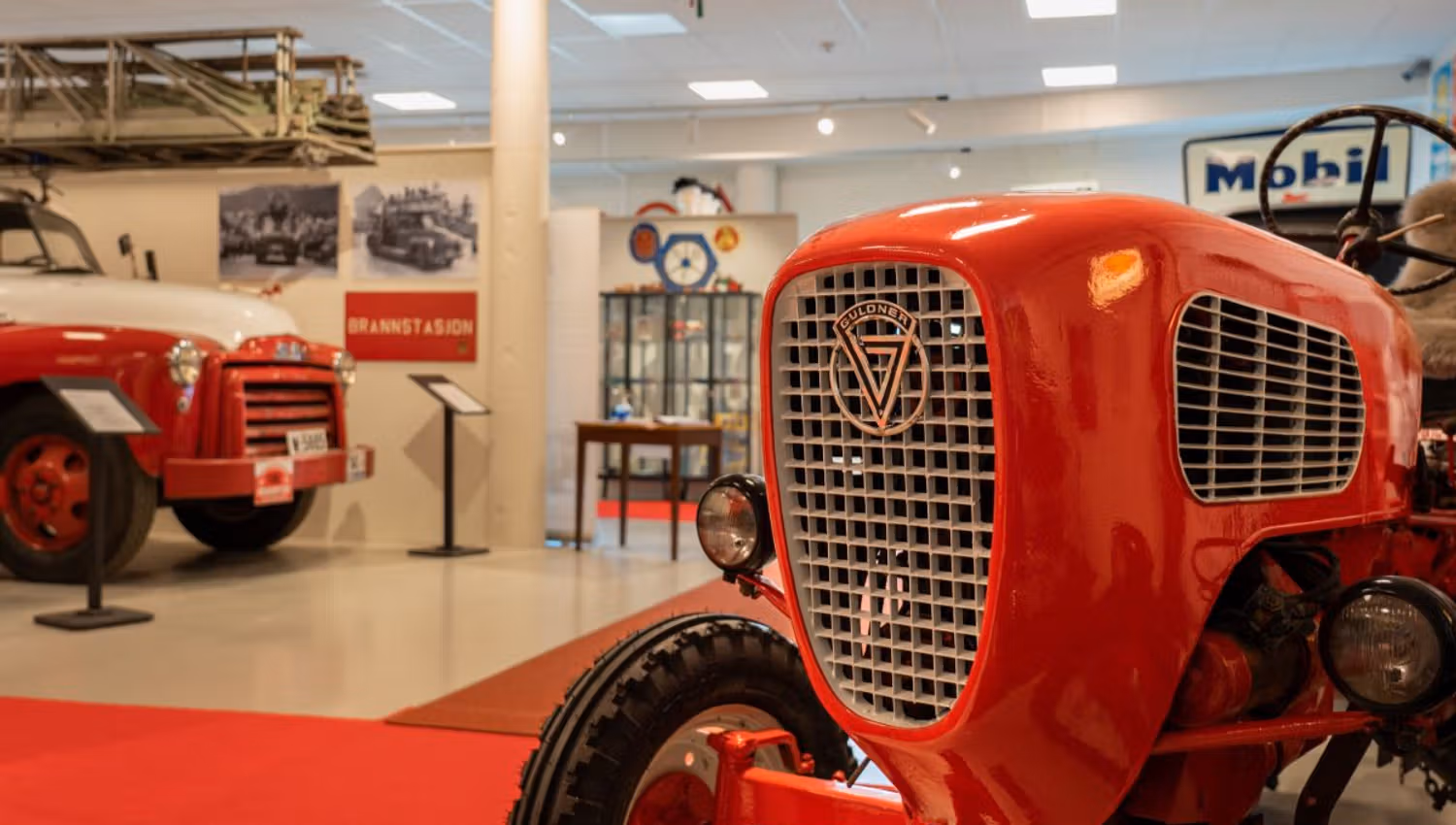 Close-up of a red vintage tractor front in an indoor museum, with another tractor and exhibits in the background.