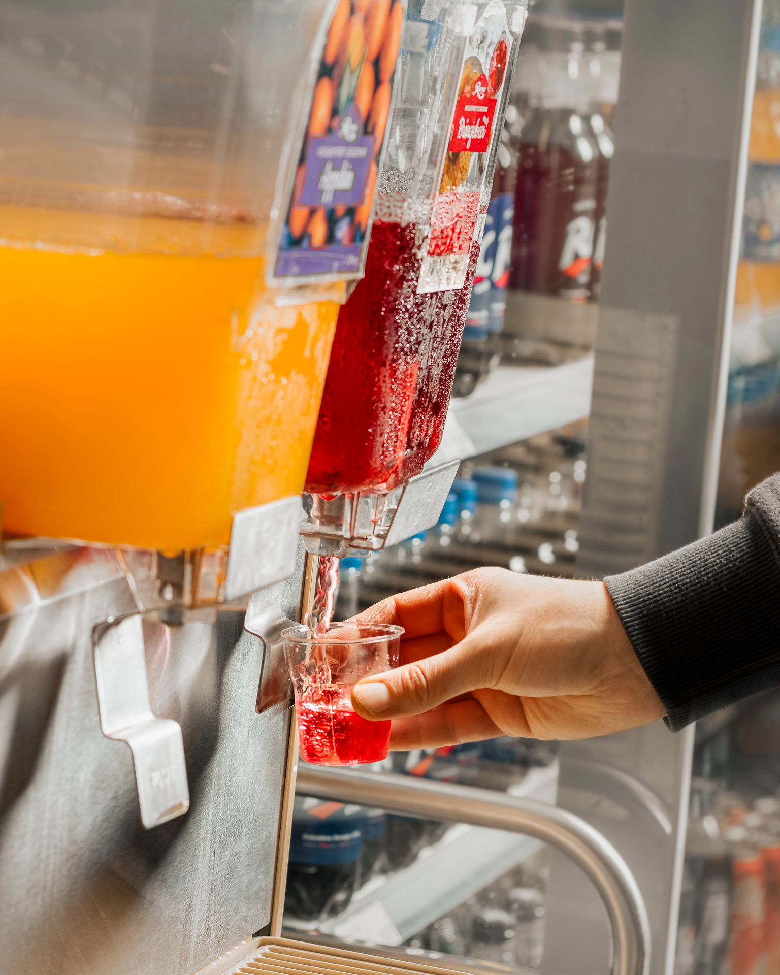 Hånd fyller et lite plastglass med rød drikke fra en saftdispenser, ved siden av en beholder med gul drikke.