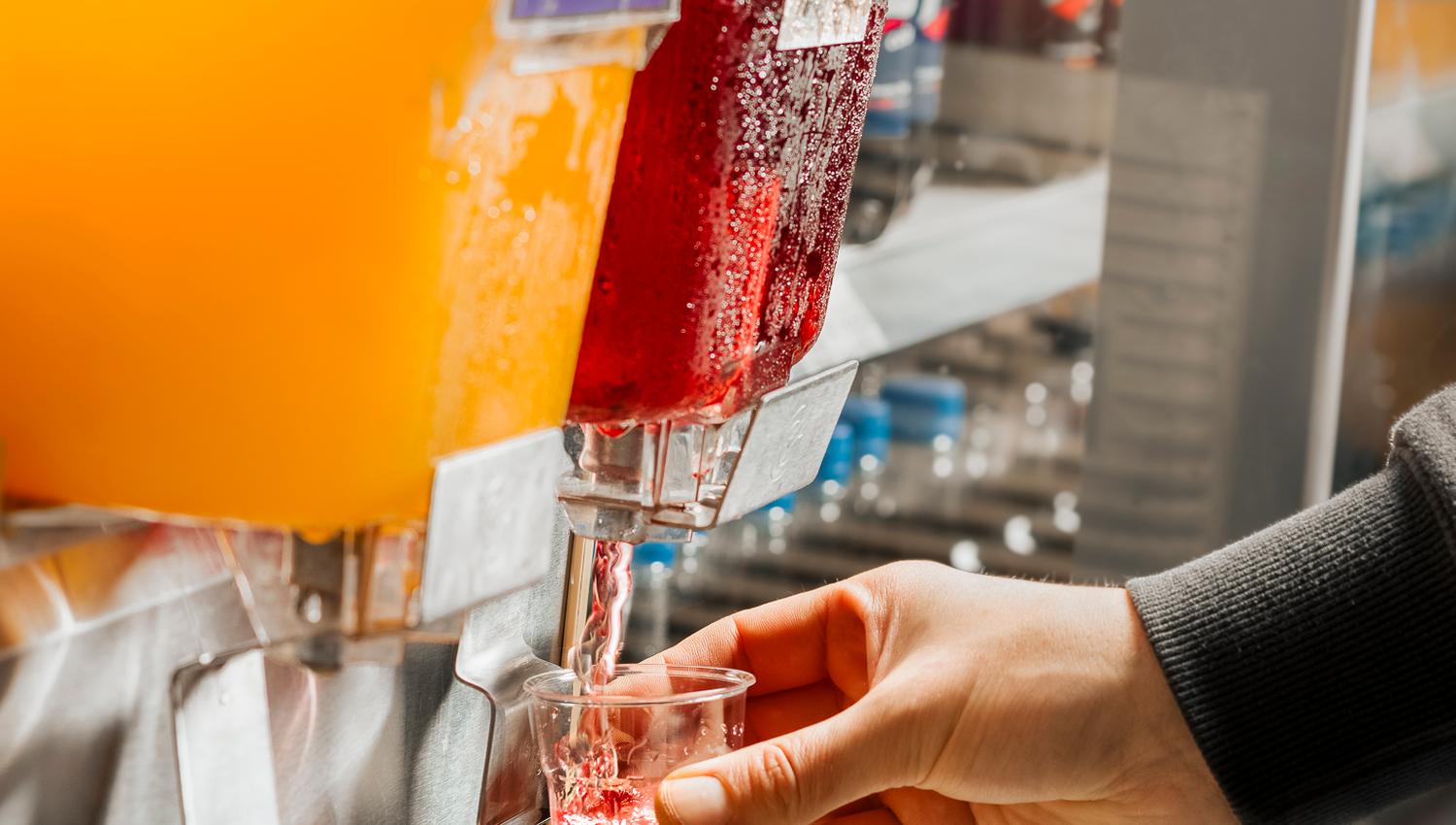 Hånd fyller et lite plastglass med rød drikke fra en saftdispenser, ved siden av en beholder med gul drikke.