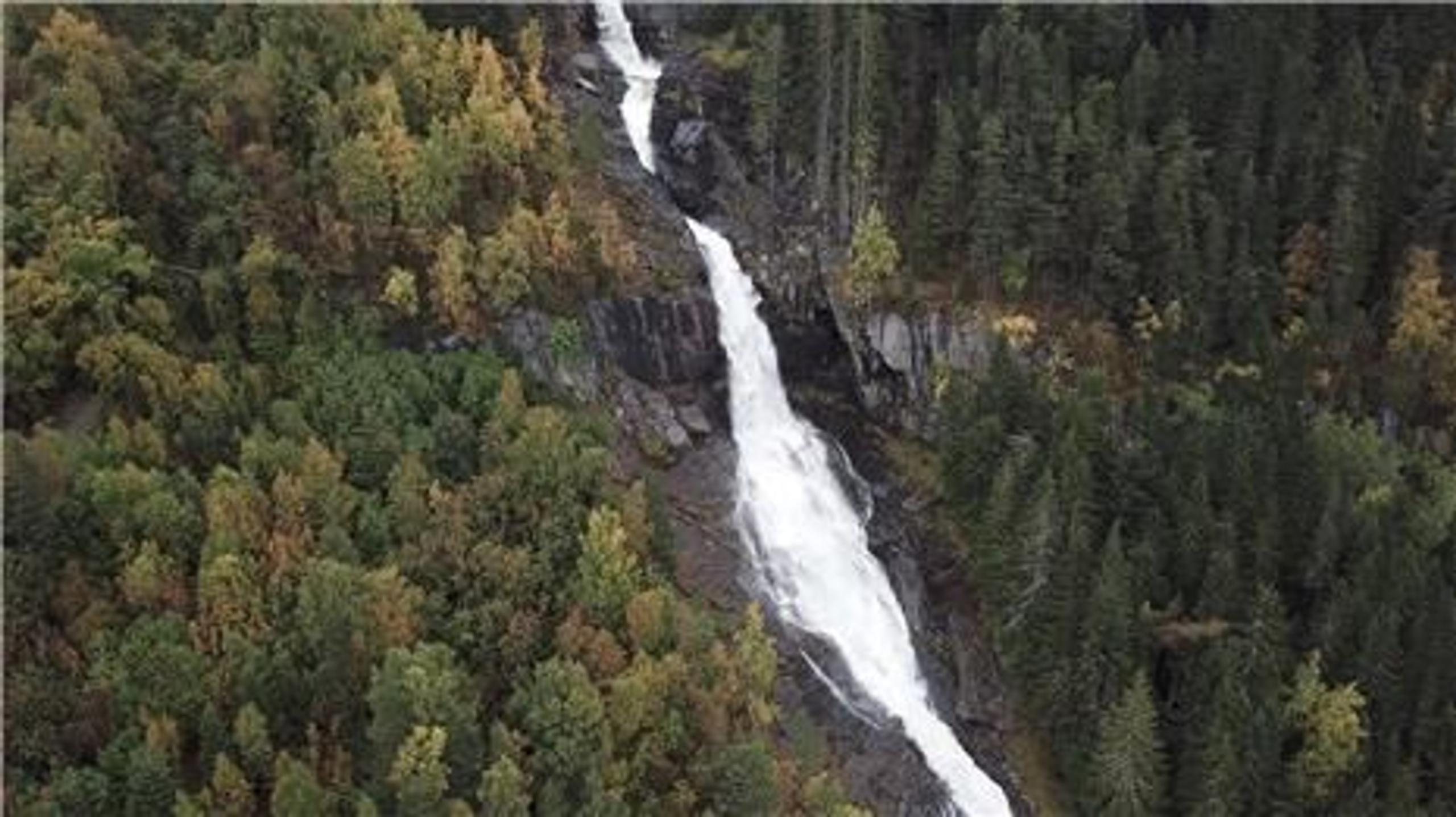 A tall white Tvinnefossen waterfall flows down a rocky gorge, surrounded by dense green and autumn-colored forest.