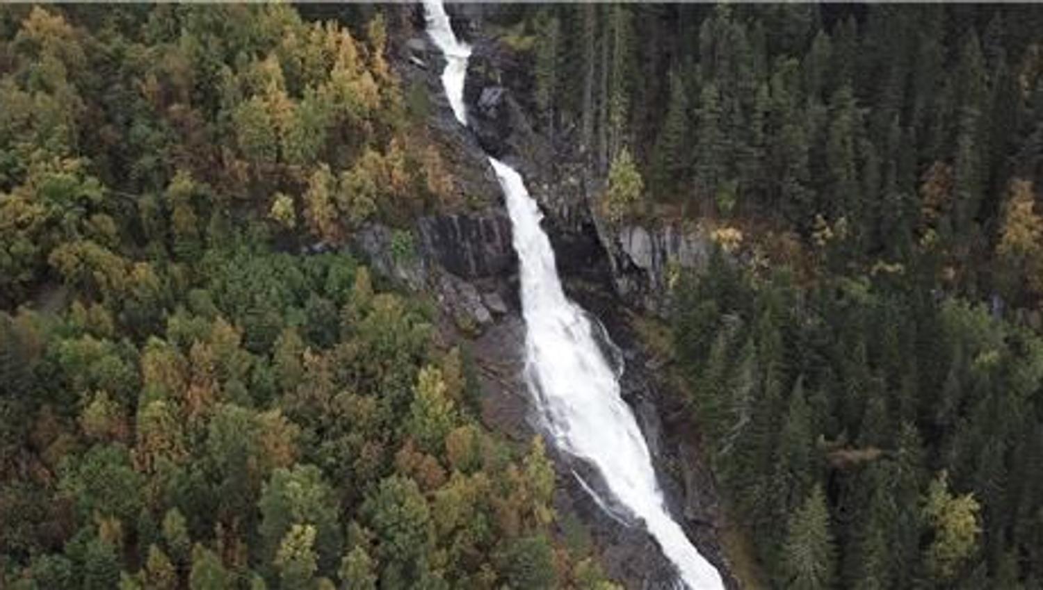 A tall white Tvinnefossen waterfall flows down a rocky gorge, surrounded by dense green and autumn-colored forest.