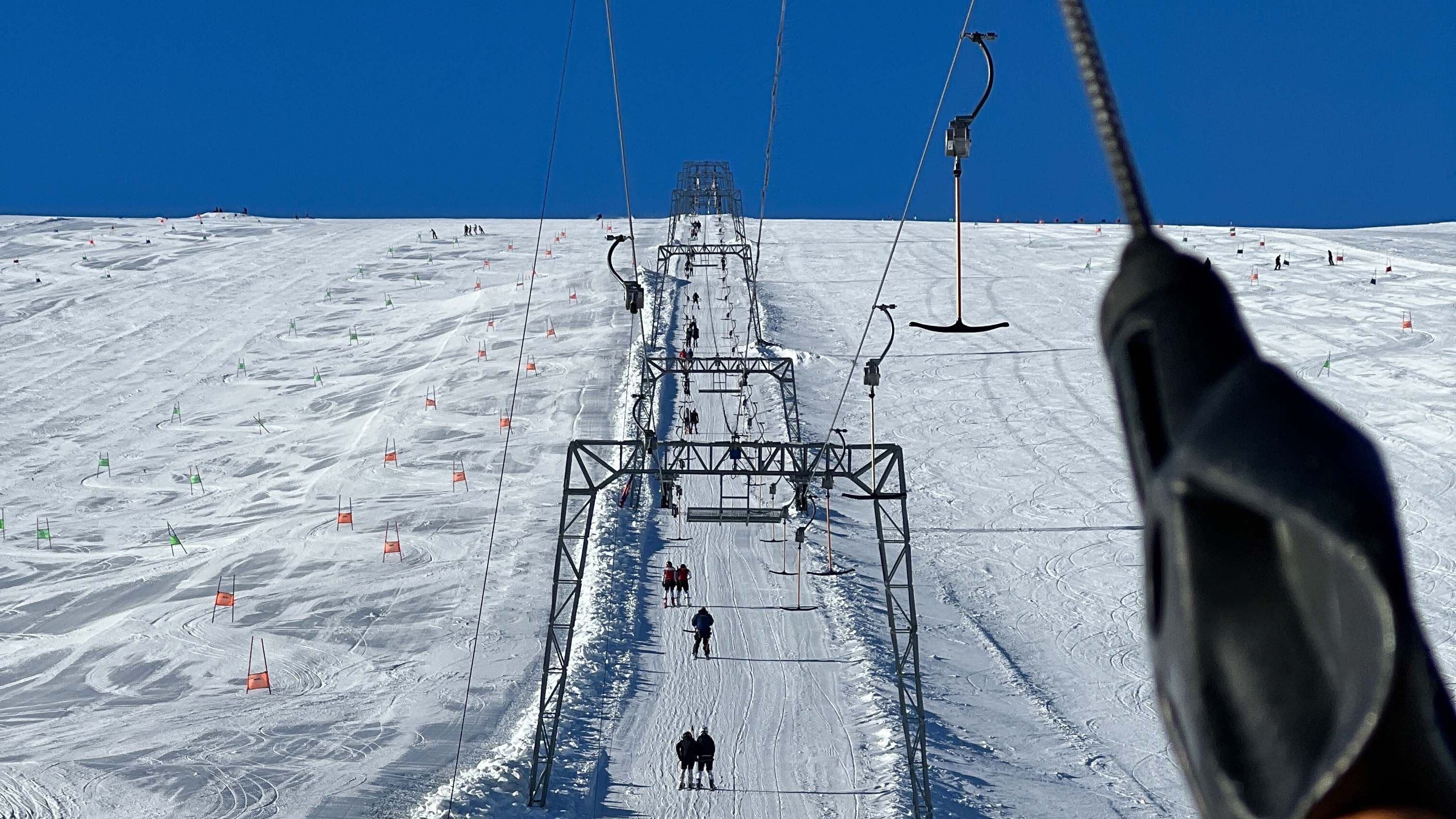 View up a snowy ski slope with a long tow lift line, skiers ascending, and orange slalom flags under blue sky..