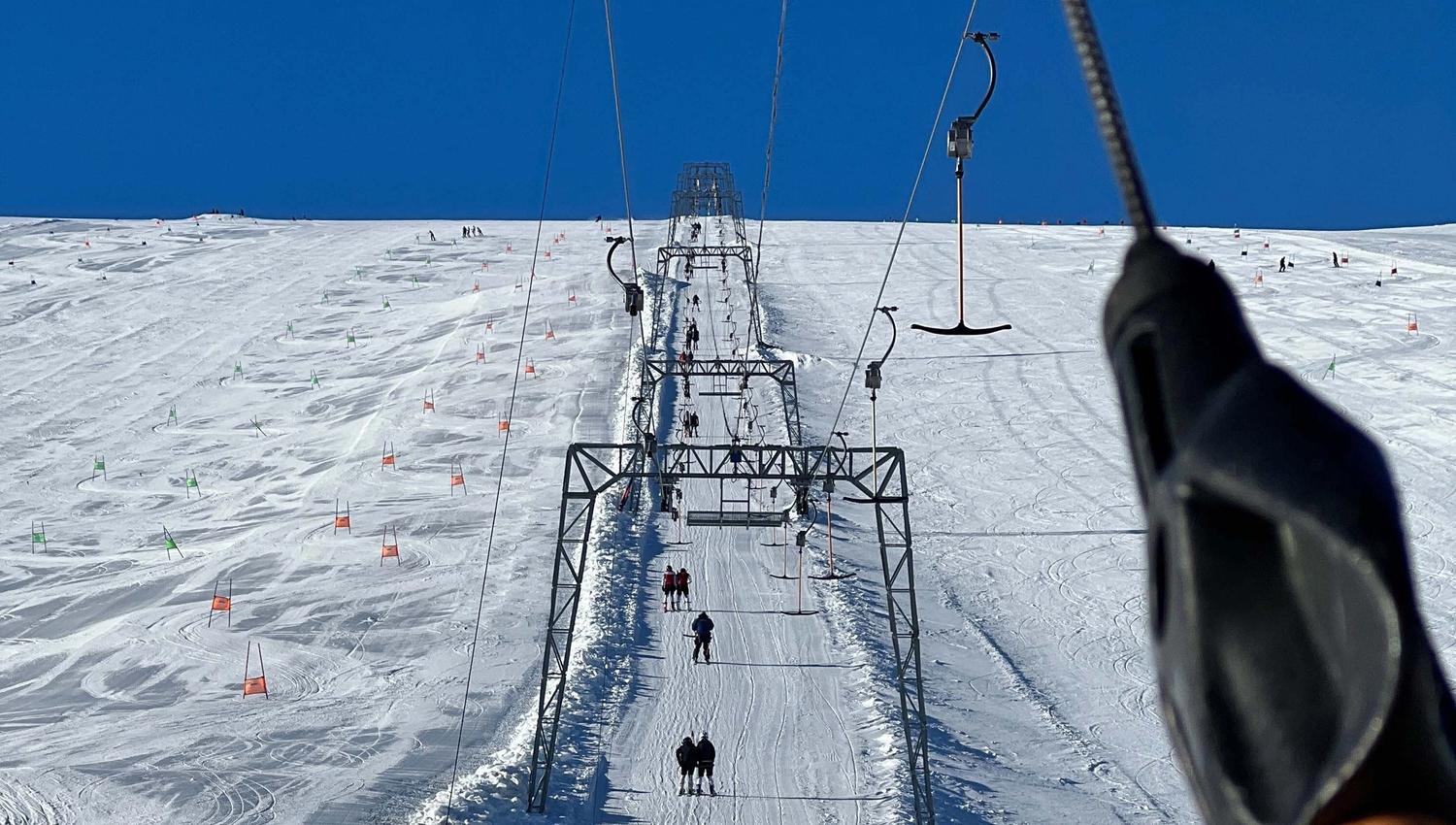 View up a snowy ski slope with a long tow lift line, skiers ascending, and orange slalom flags under blue sky..