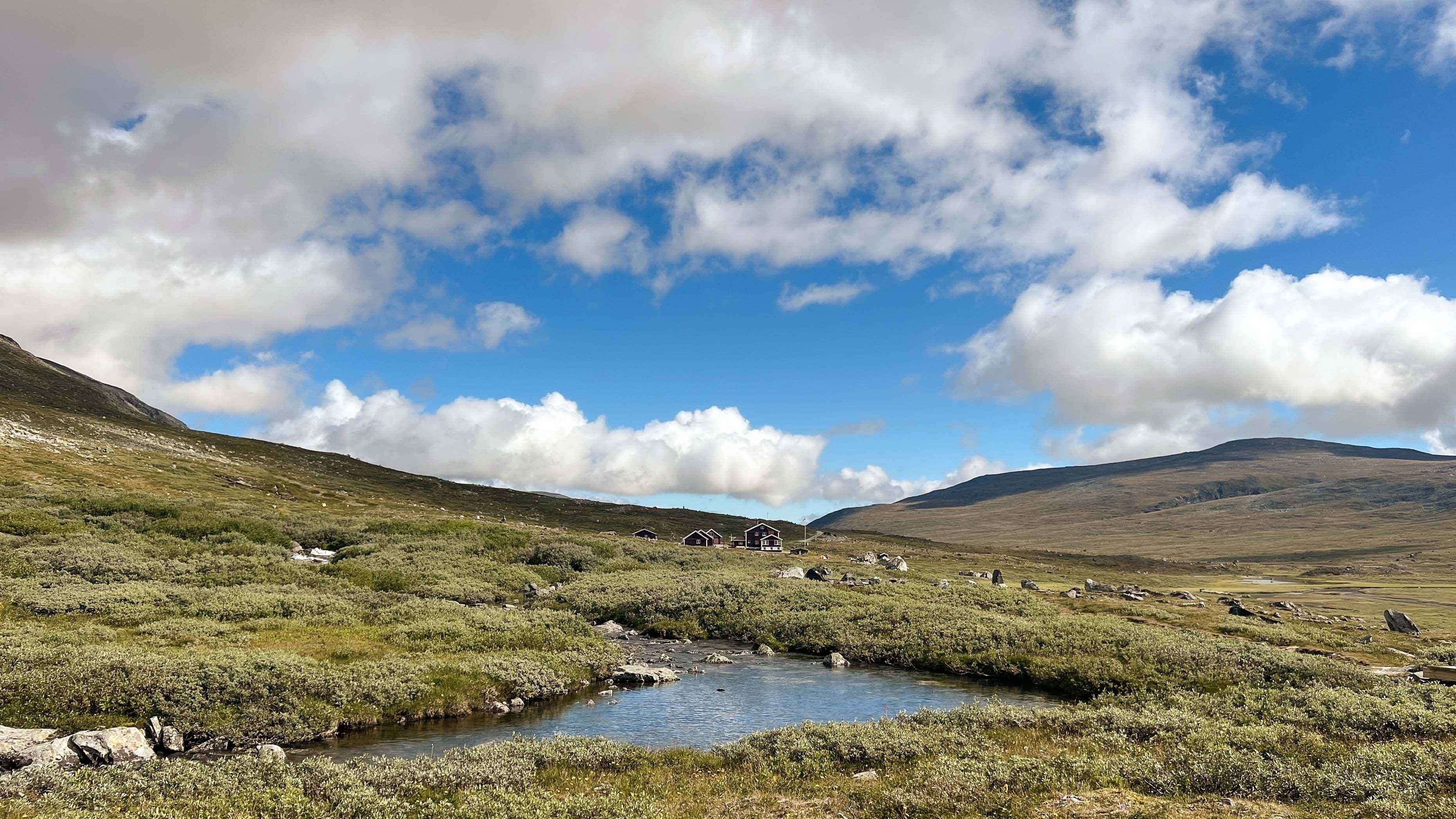 A shallow stream winds through green tundra toward small cabins, with broad hills and a blue sky with clouds..