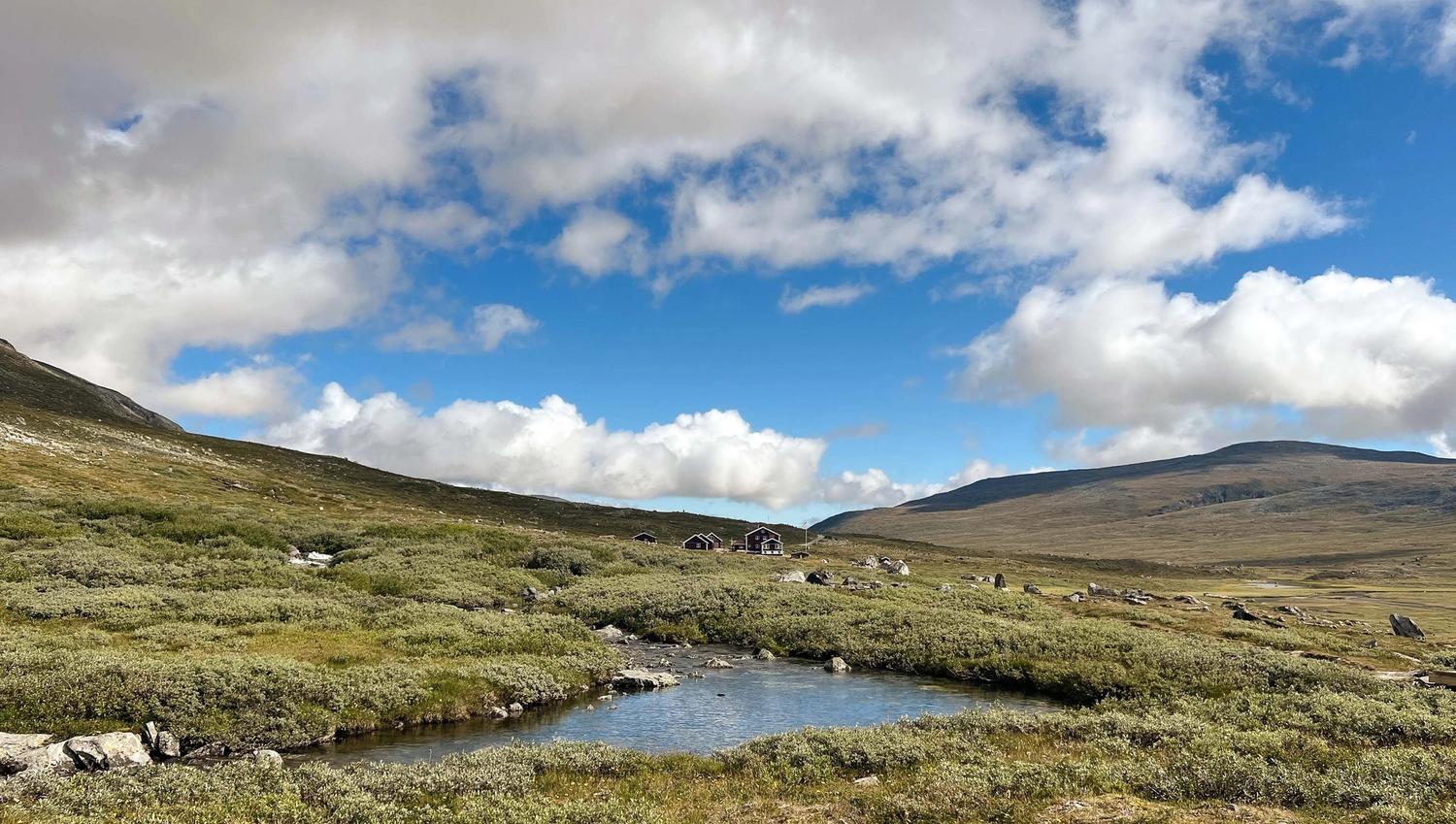 A shallow stream winds through green tundra toward small cabins, with broad hills and a blue sky with clouds..