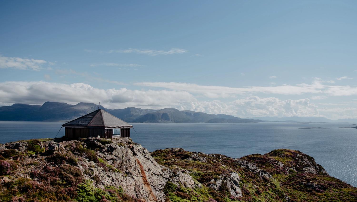 A small hut sits on a rocky hill above calm blue water, with distant mountains under a bright sky and clouds.
