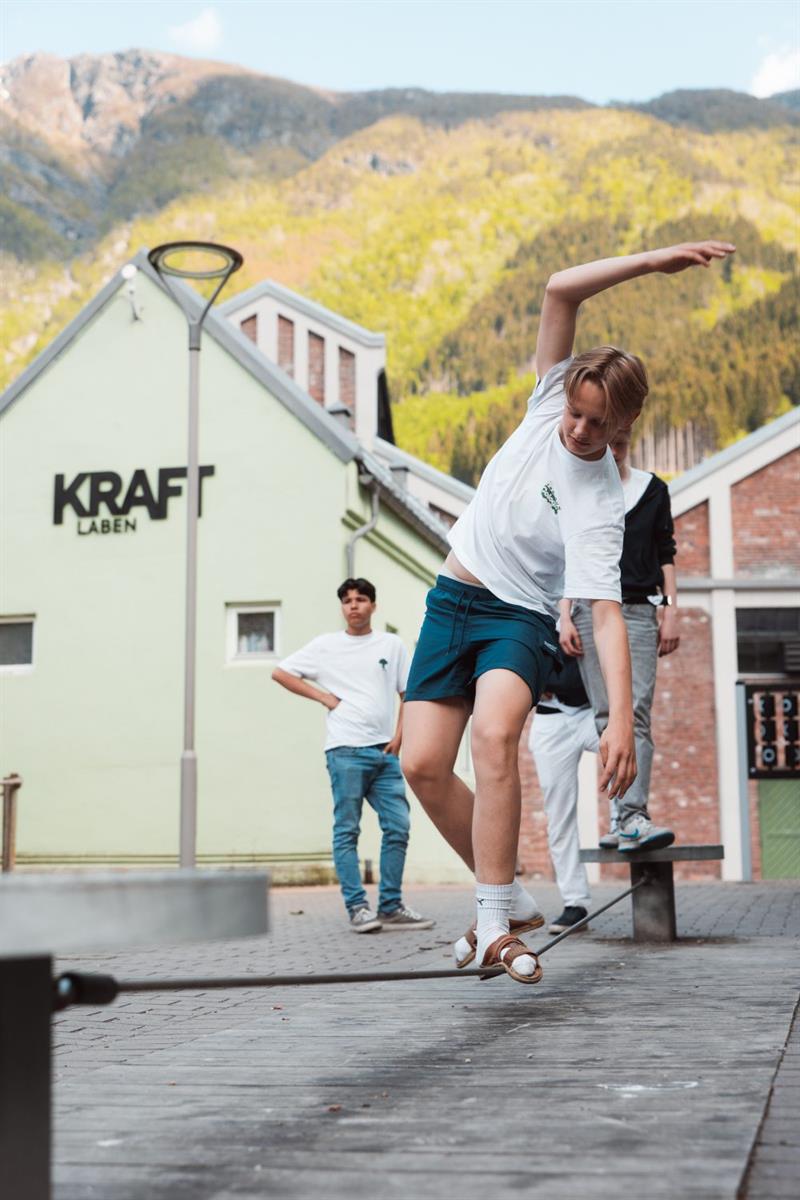 Ungdom utøver skateboard-triks på plassen utanfor KraftLaben i Odda, med Hardanger-fjella som grønt bakteppe.