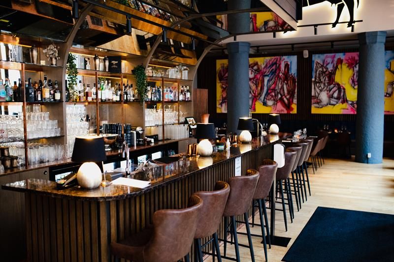 Brown bar counter and brown bar stools, shelves with bottles and glasses behind the counter, street art paintings on the wall