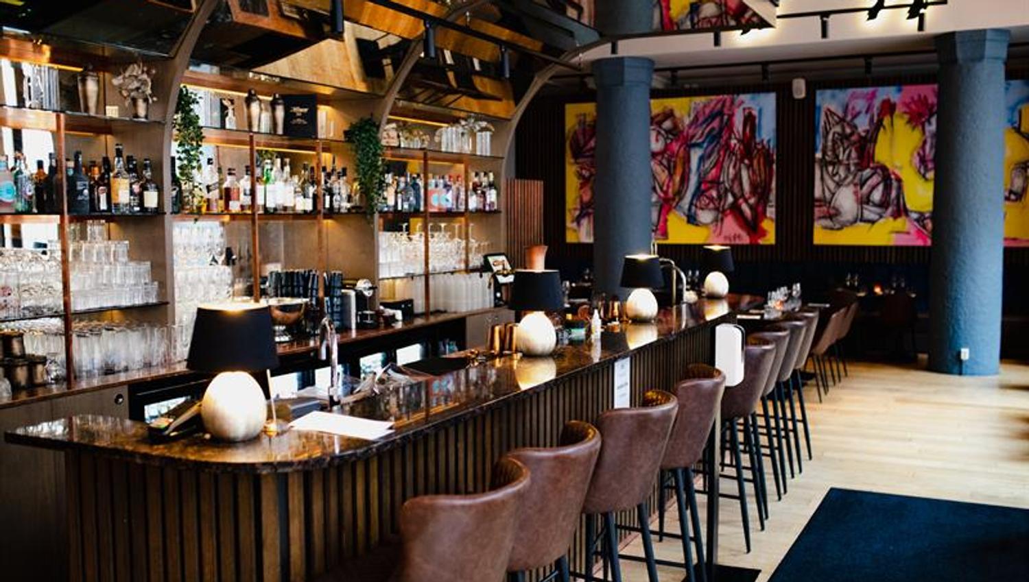 Brown bar counter and brown bar stools, shelves with bottles and glasses behind the counter, street art paintings on the wall