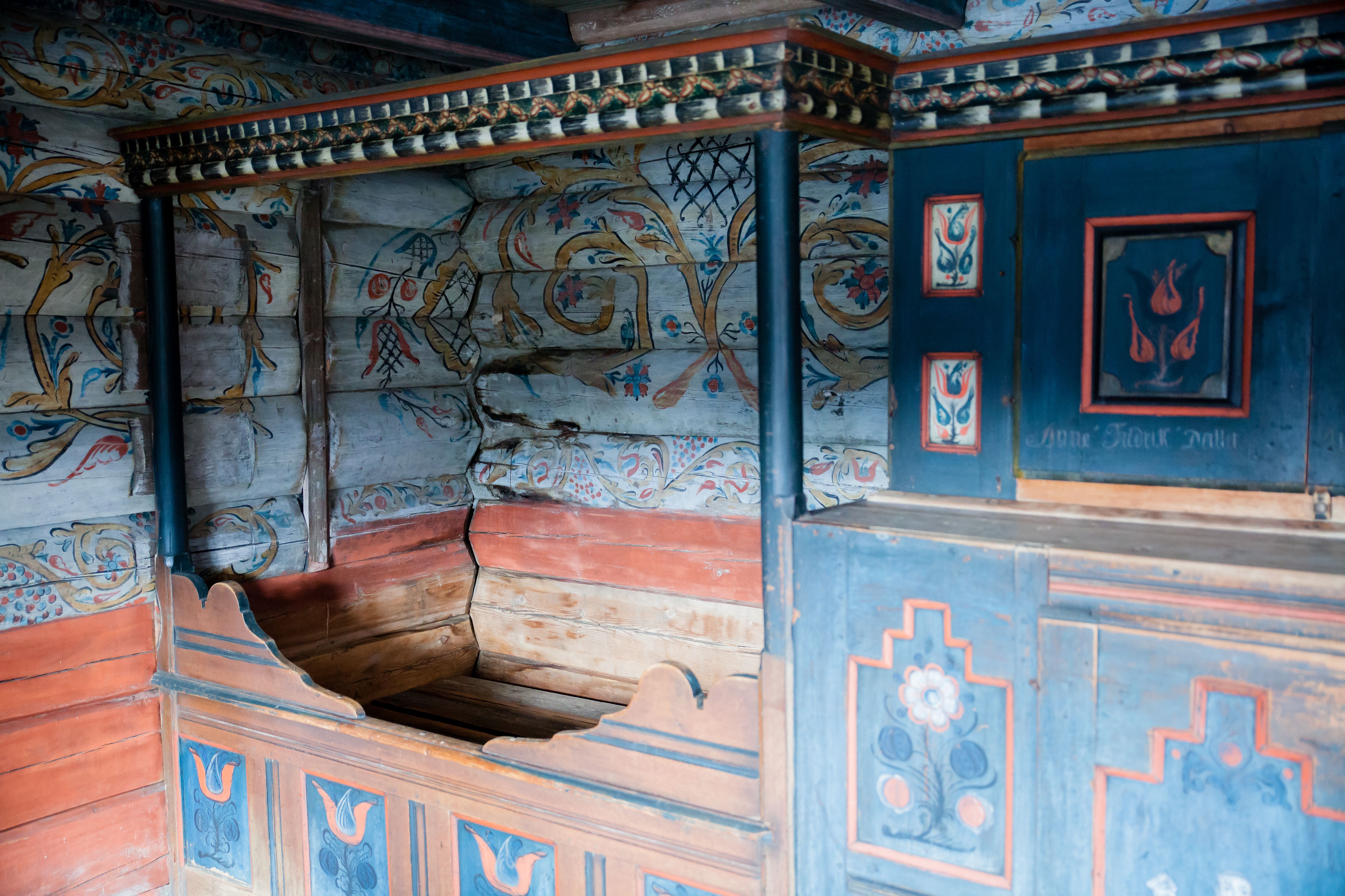 Marnardal Museum and Mjålandsstova, a wooden bed with a blue cupboard beside it. White-painted timber walls with colorful details in traditional rosem