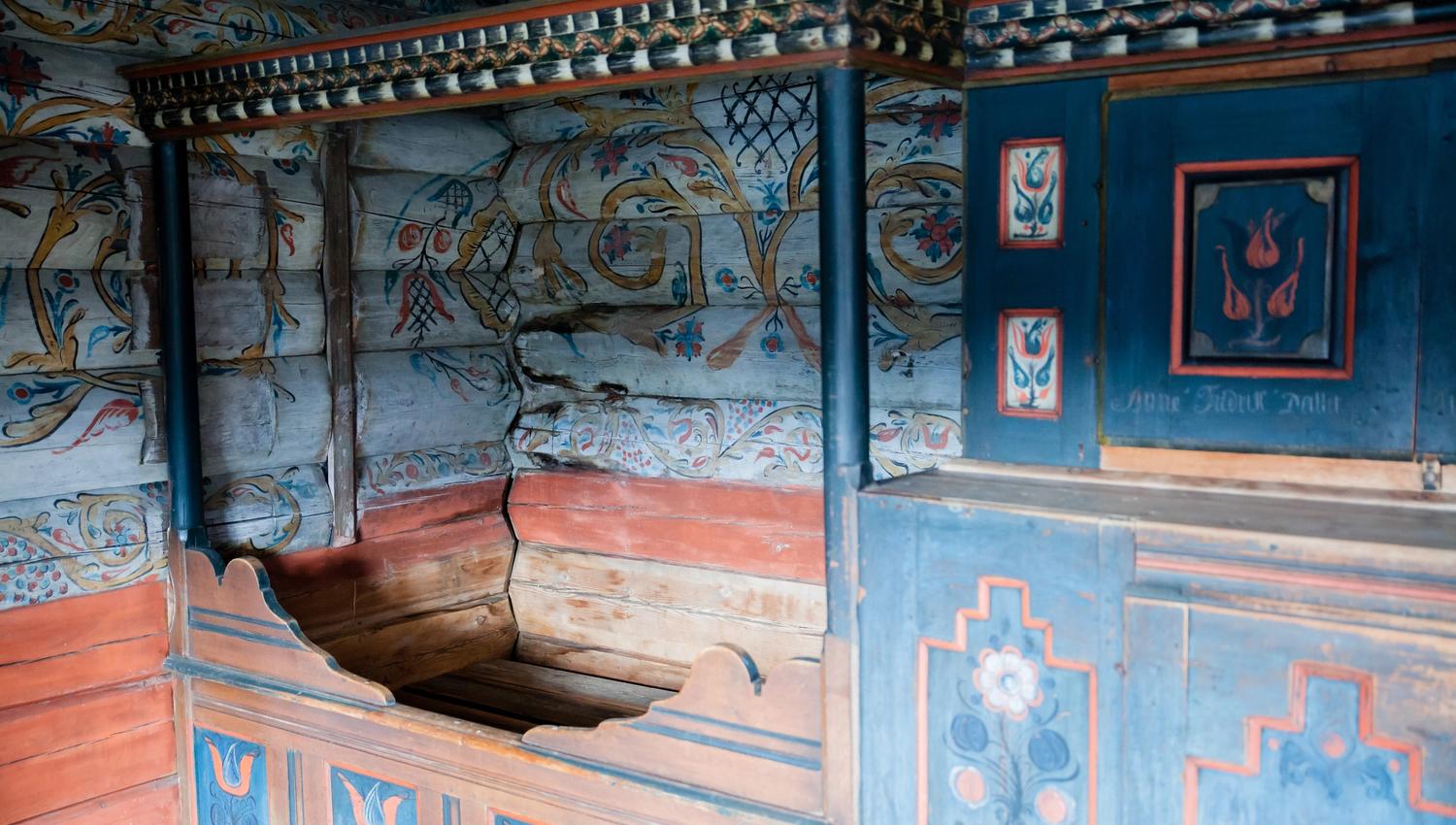 Marnardal Museum and Mjålandsstova, a wooden bed with a blue cupboard beside it. White-painted timber walls with colorful details in traditional rosem