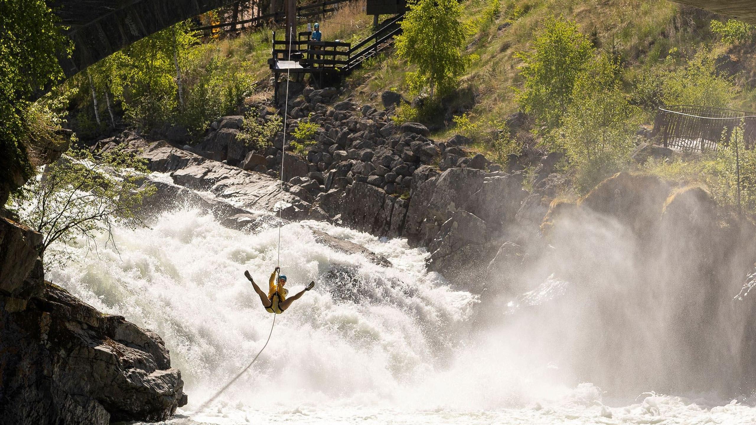 A person wearing a helmet and harness swings on a rope above foaming river rapids beside rocky cliffs and trees..