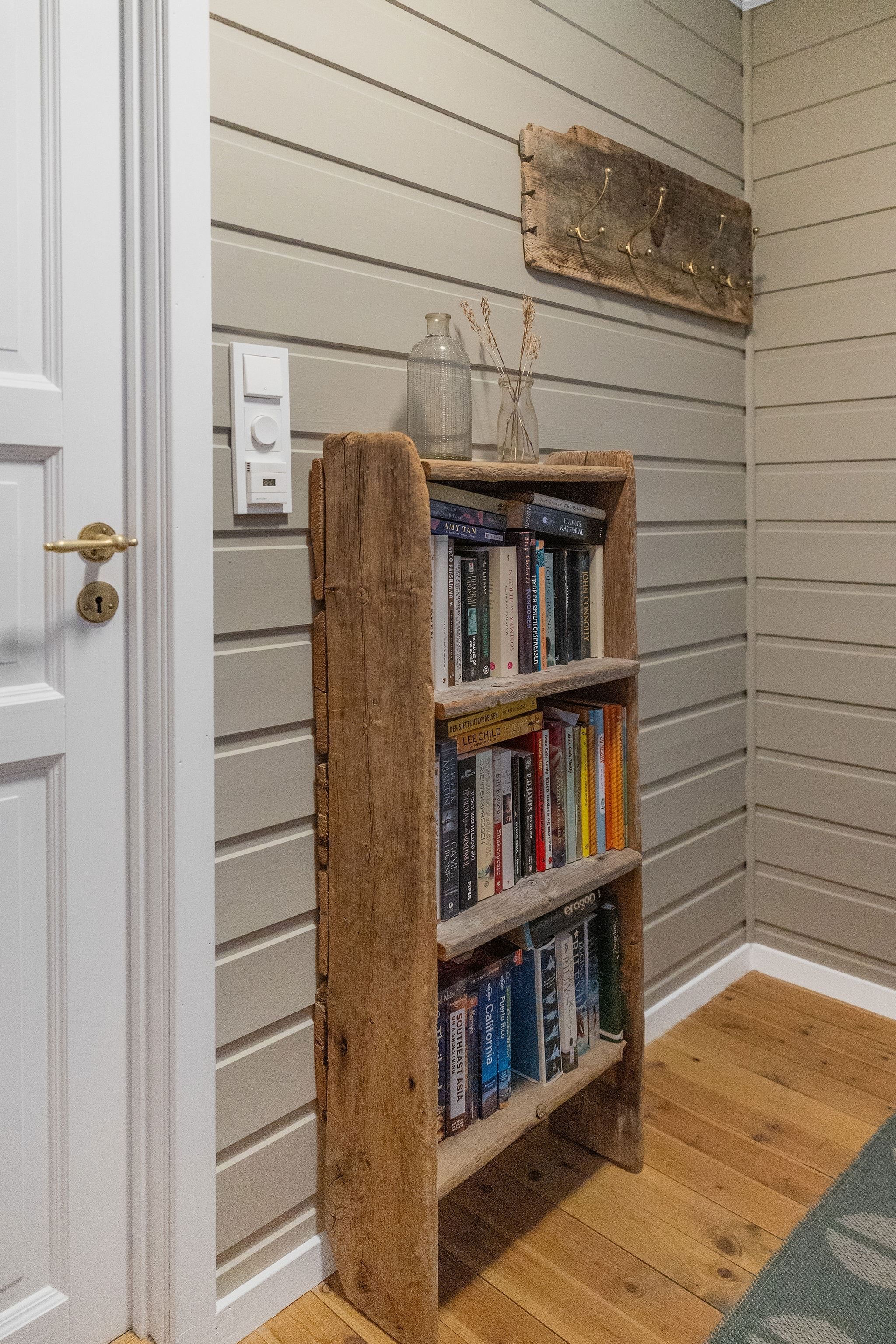 Wooden bookshelf filled with books stands by a white door on a gray paneled wall with coat hooks above at the Seahouse at Hornelen Panorama in Bremanger..