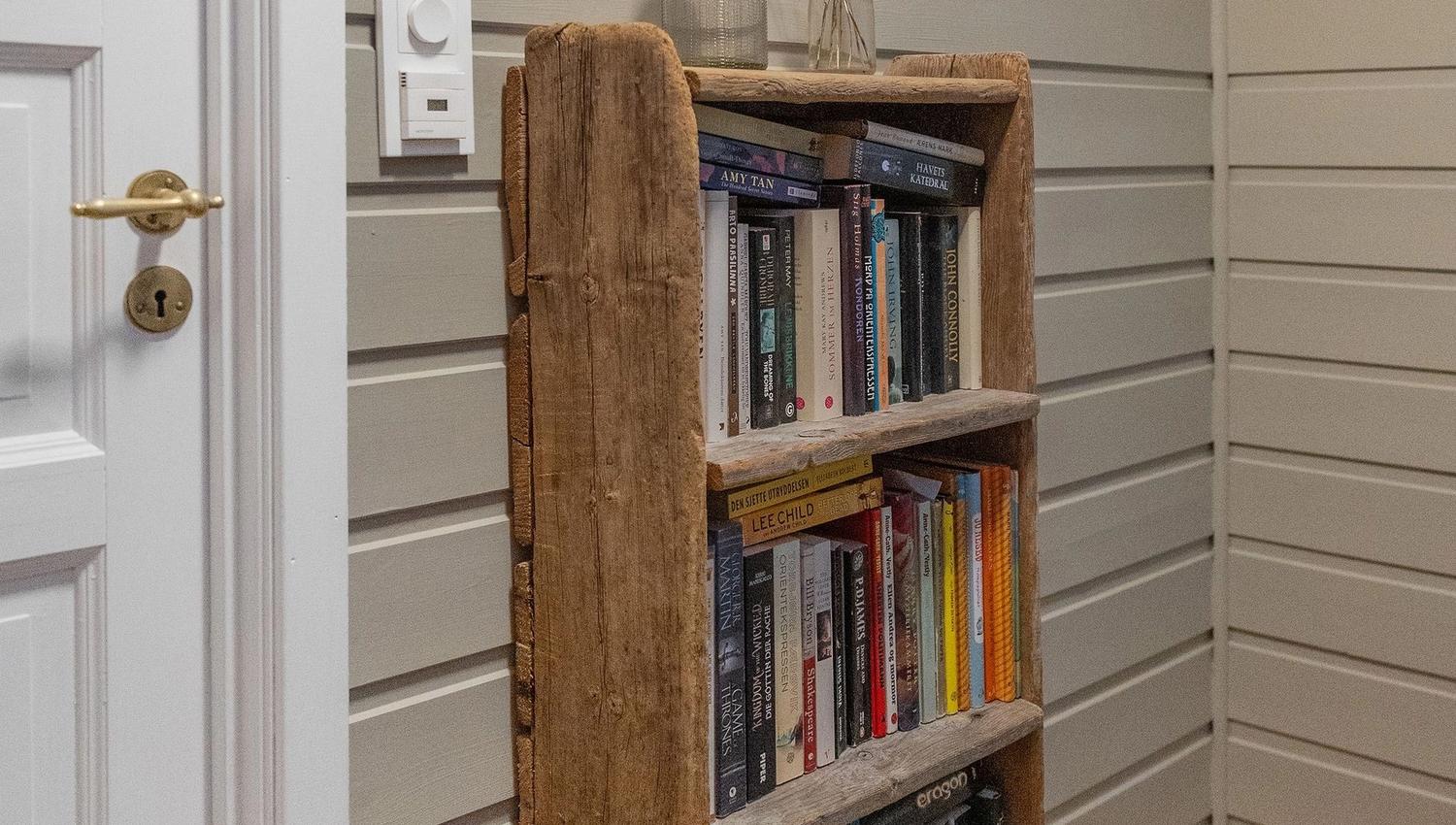 Wooden bookshelf filled with books stands by a white door on a gray paneled wall with coat hooks above at the Seahouse at Hornelen Panorama in Bremanger..