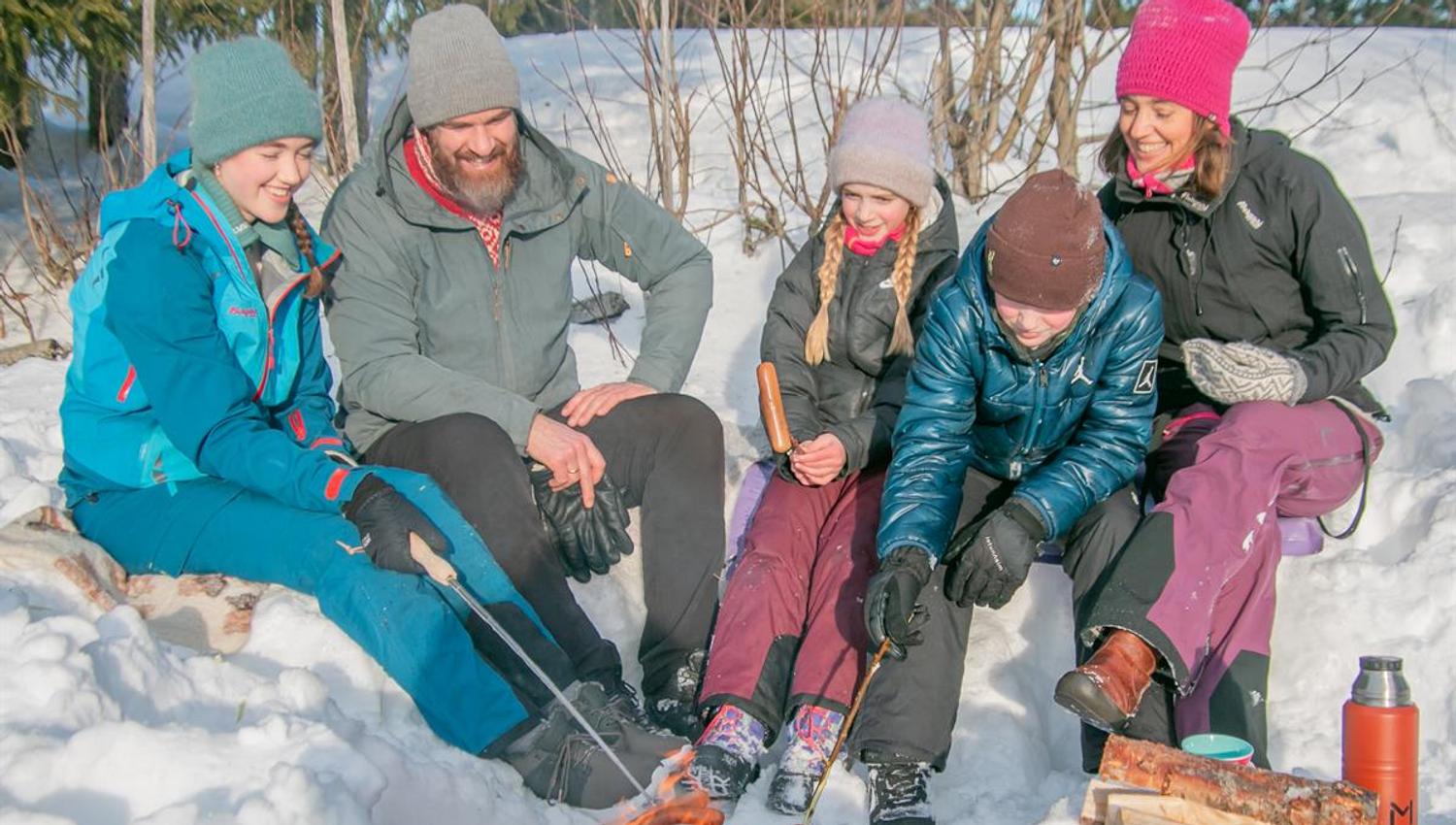 Familie sitter i snøen og varmer drikke ved et bål på en solrik vinterdag.