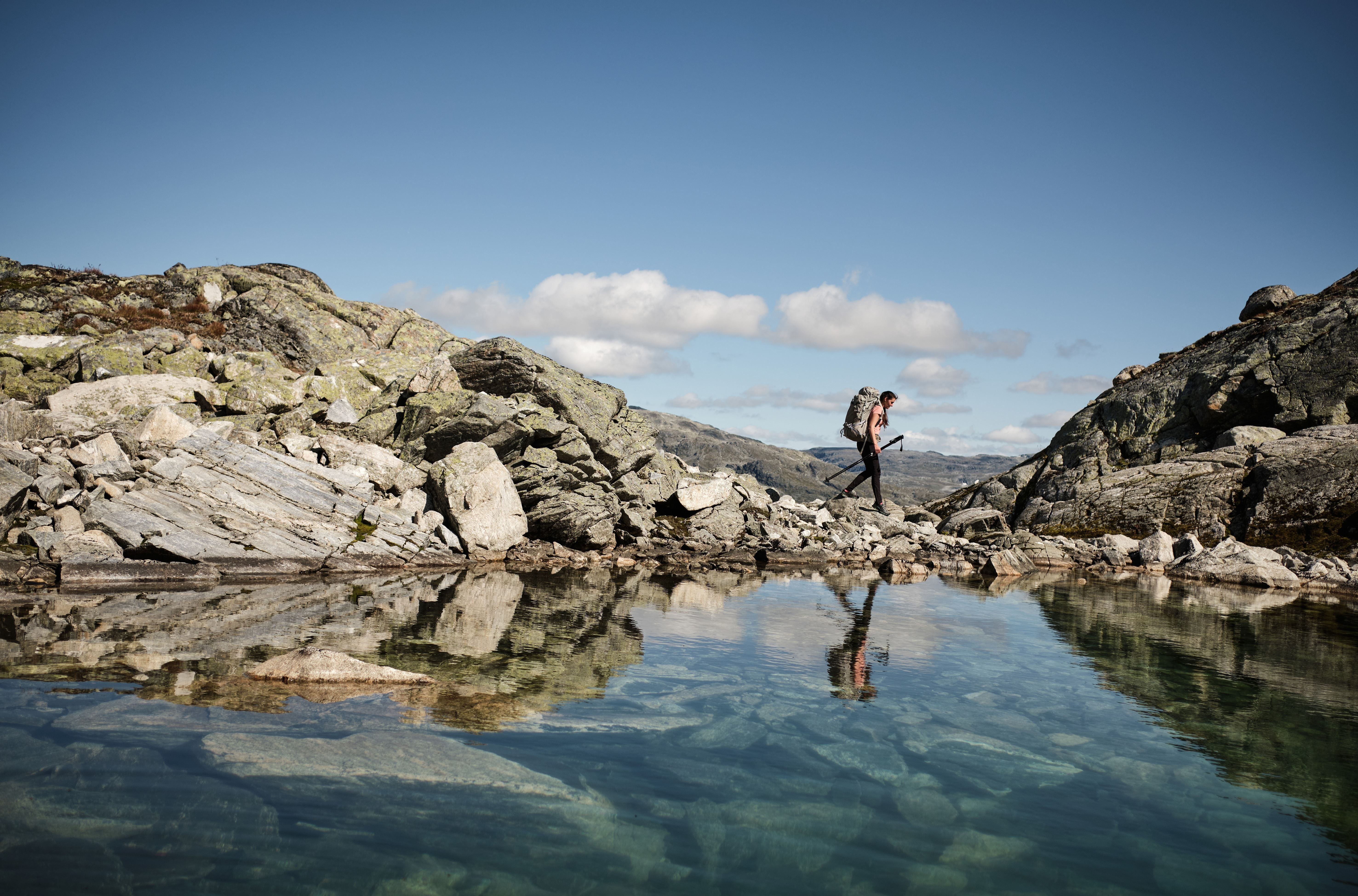 Ein person går langs eit fjellvatn. I vatnet vert både berget, mannen og den klare blå himmelen reflektert. 