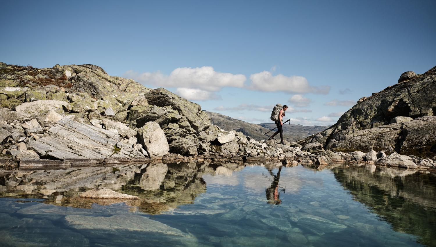 Ein person går langs eit fjellvatn. I vatnet vert både berget, mannen og den klare blå himmelen reflektert.