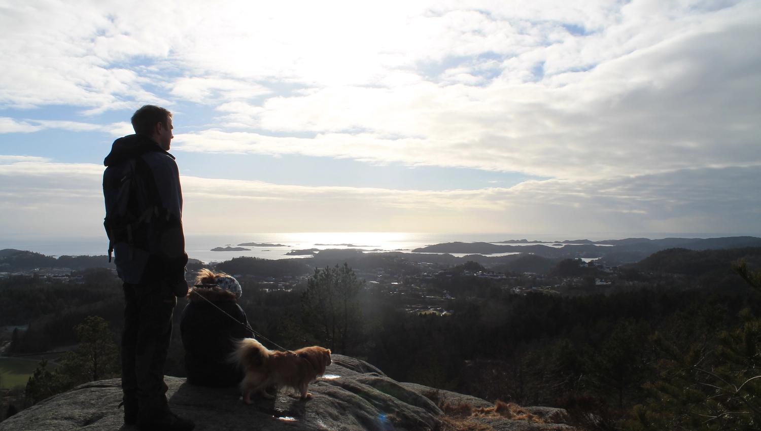 En mann som står og en dame som sitter ved en liten hund (tibetansk spaniel) ser utover mot husene, fjellet og havet. Sol bak skyene på himmelen