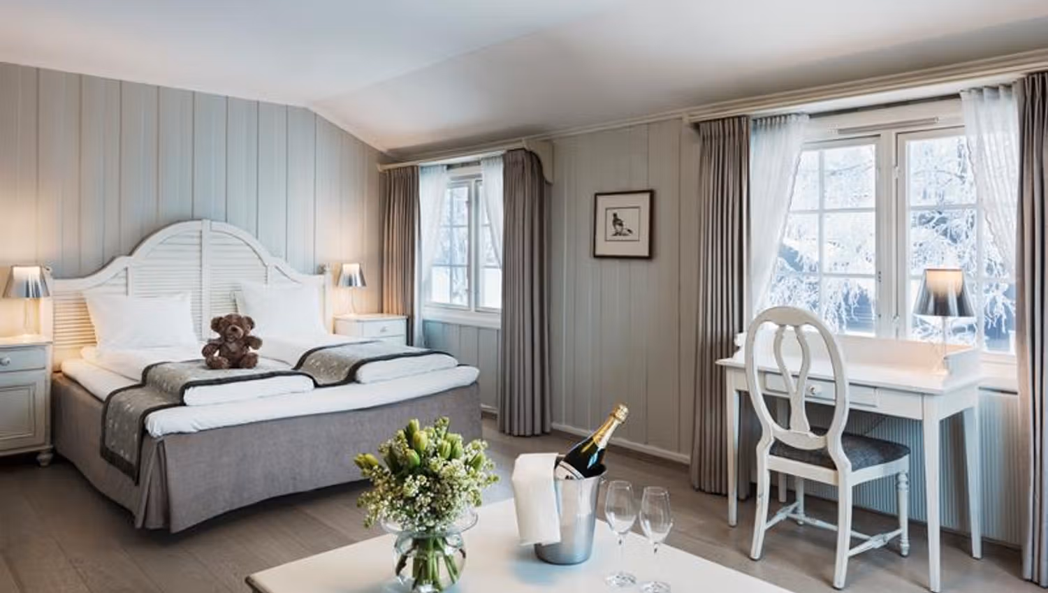 Double room in tones of grey and white. Desk, teddy bear on the bed, champagne and flowers on the table. Photo.