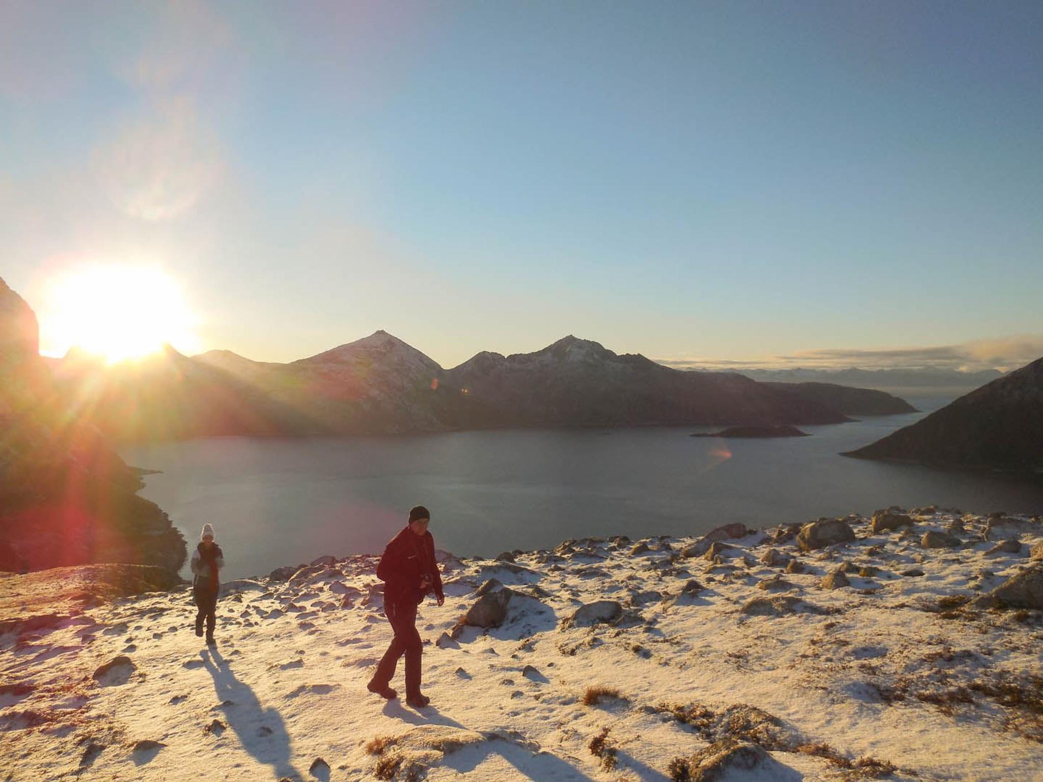 To personer går i snødekt fjellterreng med solnedgang og vann i bakgrunnen.