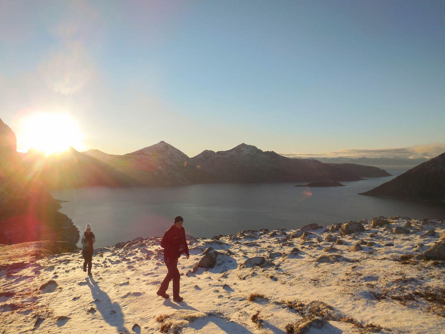 To personer går i snødekt fjellterreng med solnedgang og vann i bakgrunnen.