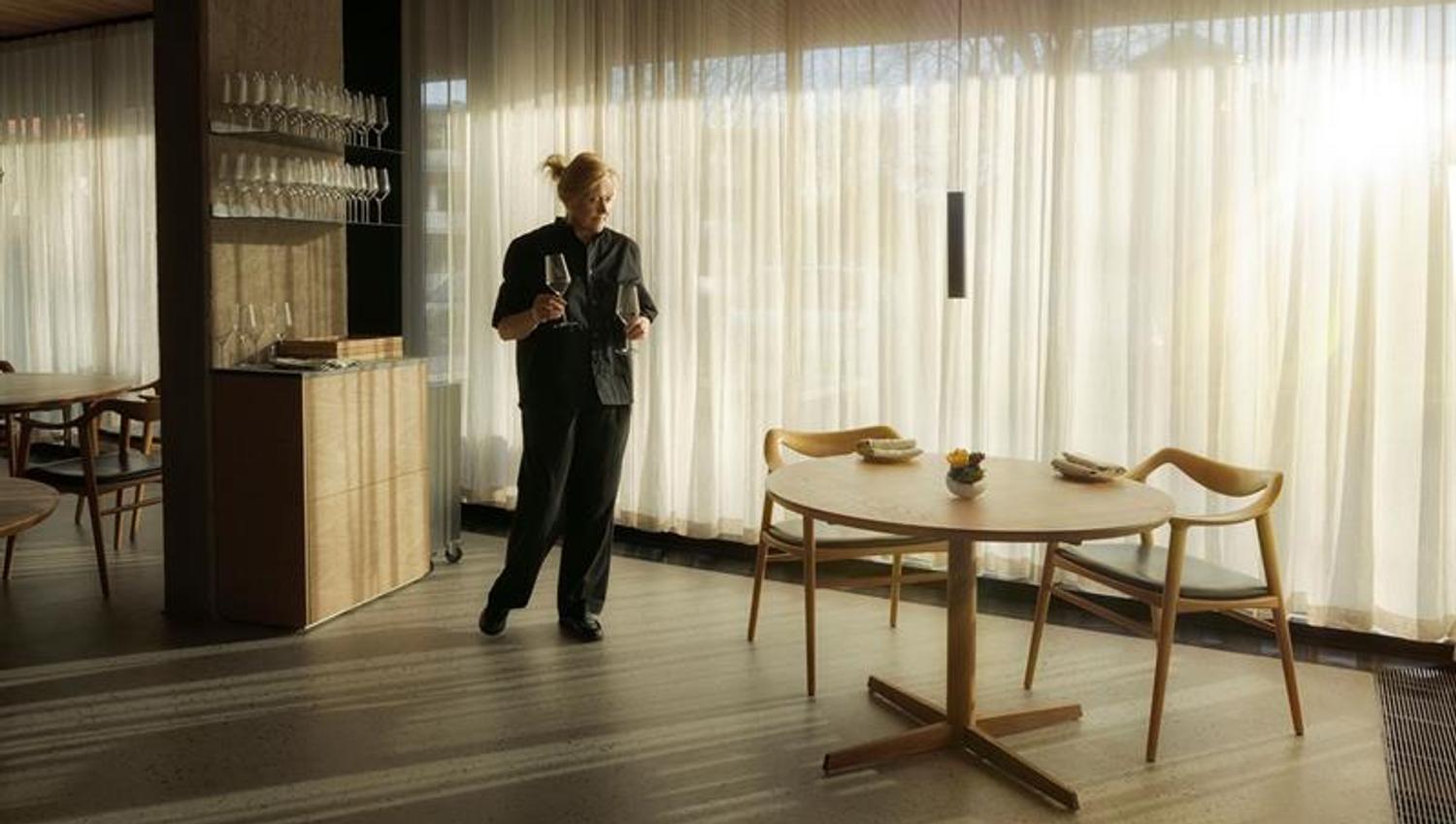 Dame som står med to vinglass foran et stort panoramavindu med lyse gardiner