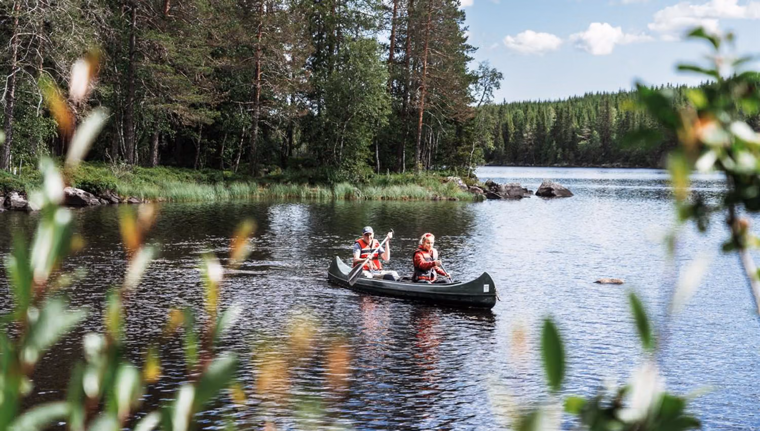 Two people canoeing on a calm forest lake, viewed through blurred reeds in the foreground.