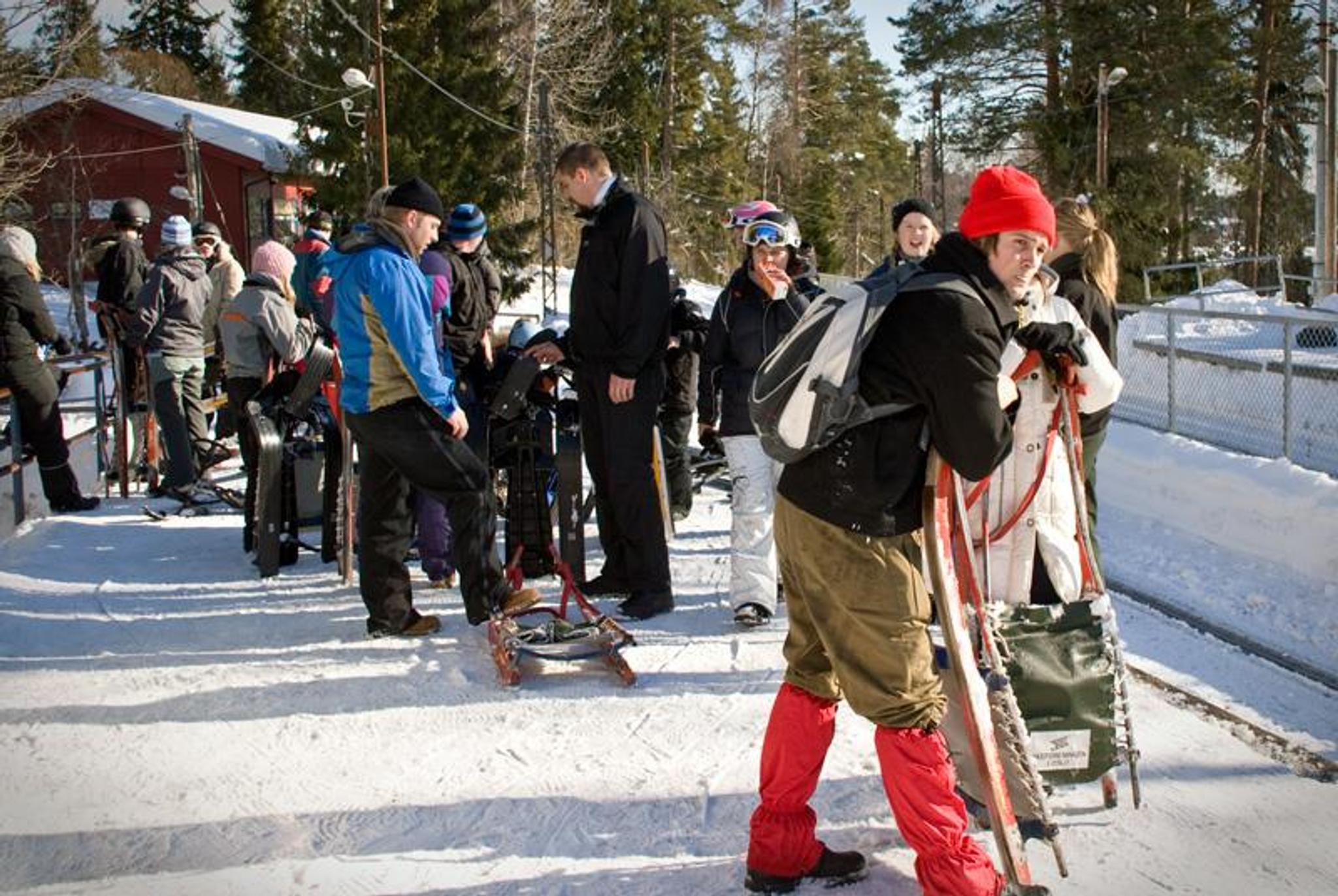 Mennesker med ski og akebrett på snødekt bakke, med trær i bakgrunnen.
