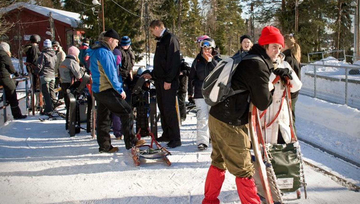 Mennesker med ski og akebrett på snødekt bakke, med trær i bakgrunnen.