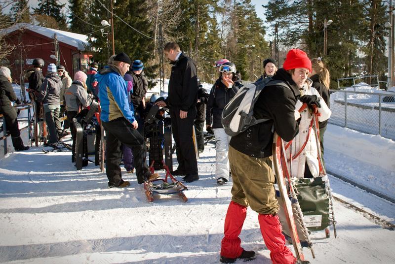 Mennesker med ski og akebrett på snødekt bakke, med trær i bakgrunnen.