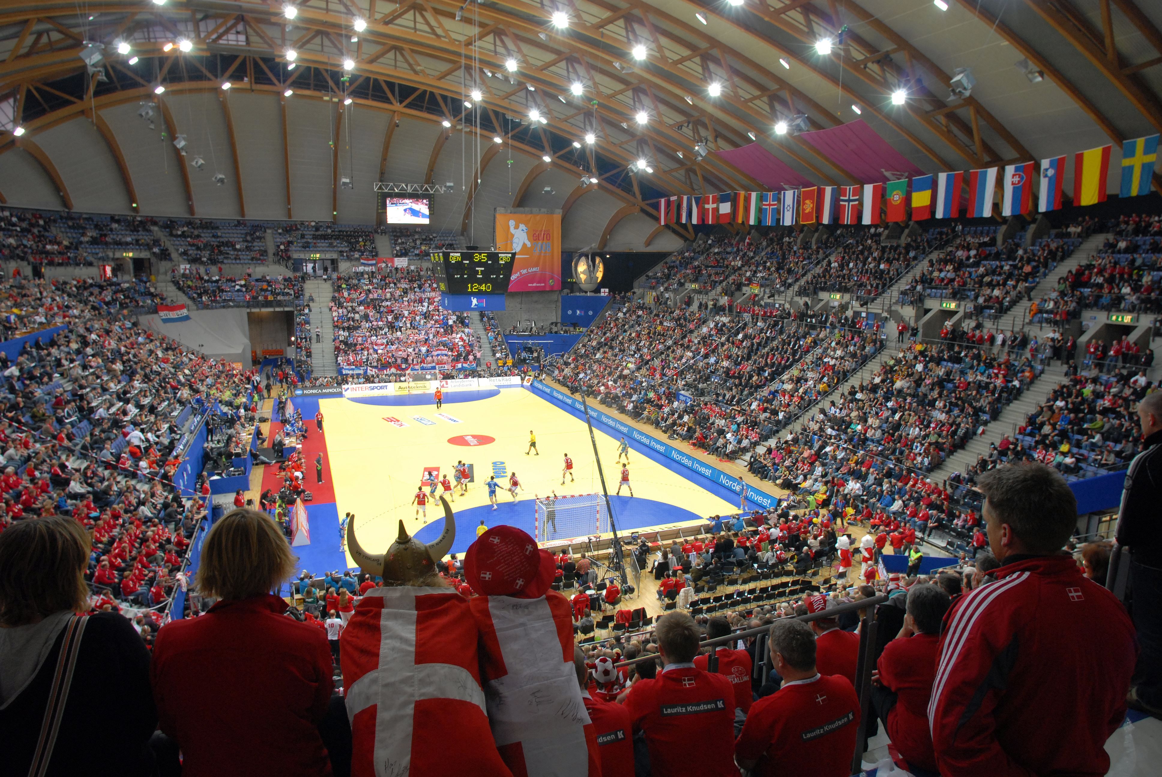 Fullsatt innendørs håndballarena sett fra tribunen, med kamp på banen, flaggrekke og lystavle i taket..