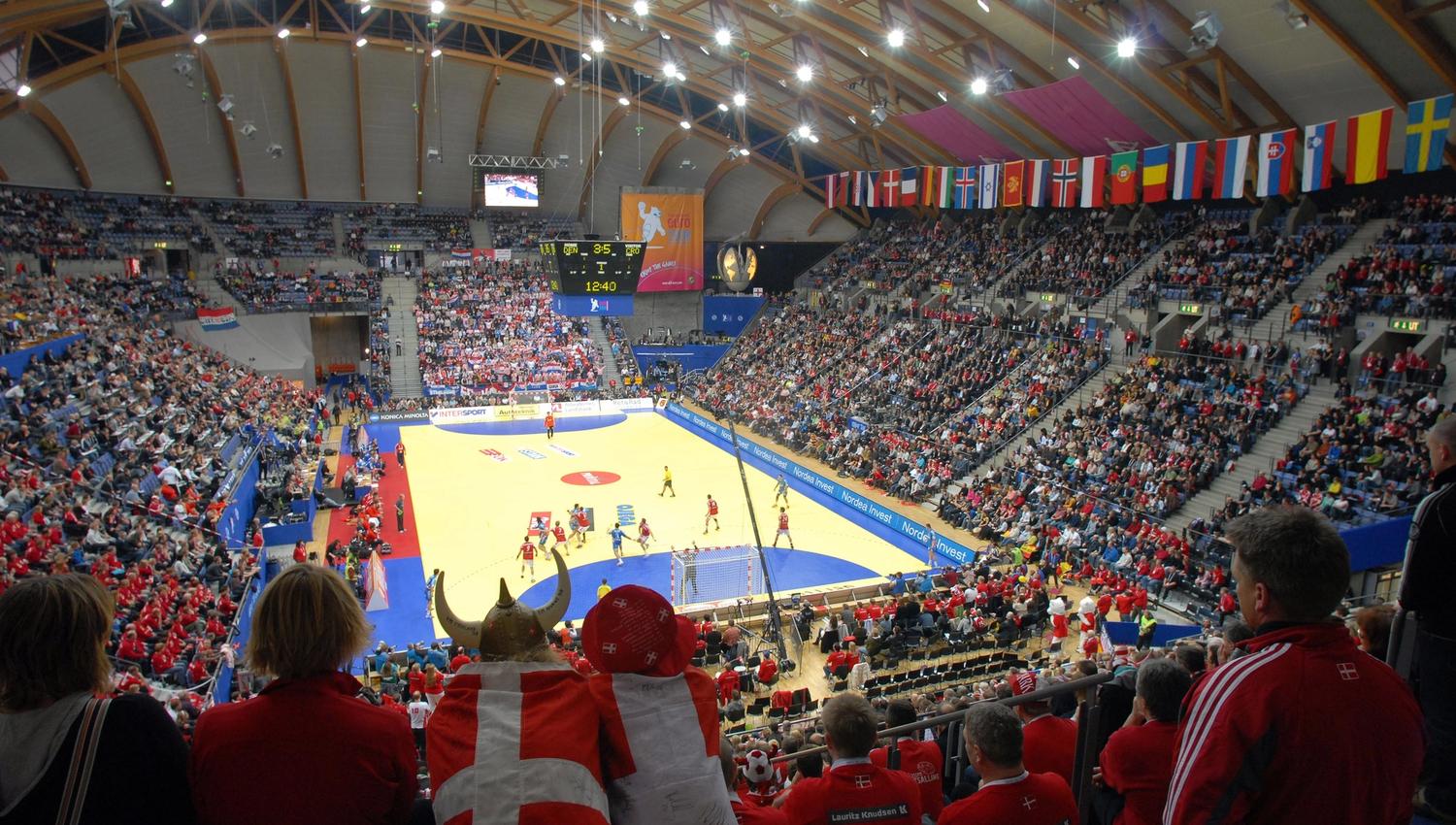 Fullsatt innendørs håndballarena sett fra tribunen, med kamp på banen, flaggrekke og lystavle i taket..